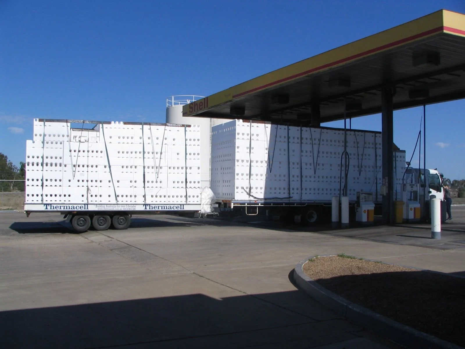 A gas station with trucks carrying large white foam blocks, a Shell sign overhead, and a person near the pumps.