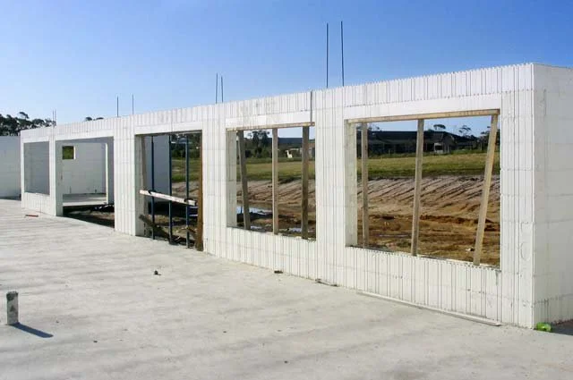Construction site with unfinished white framed building with window openings, on a concrete foundation with dirt and construction materials nearby.