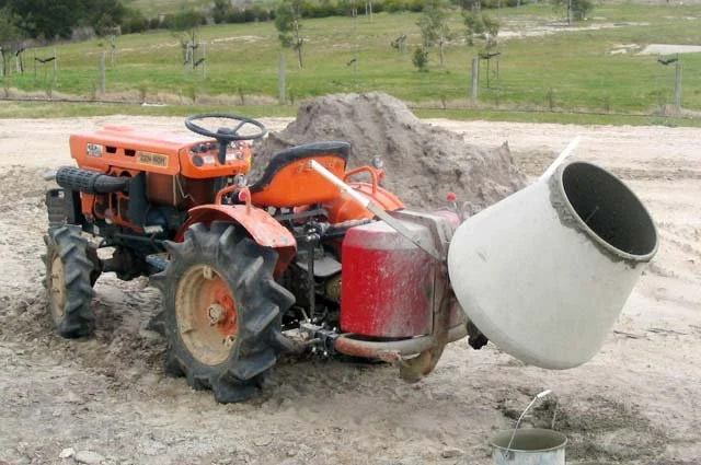 Construction site with an orange tractor attaching a large concrete mixer drum to the back.