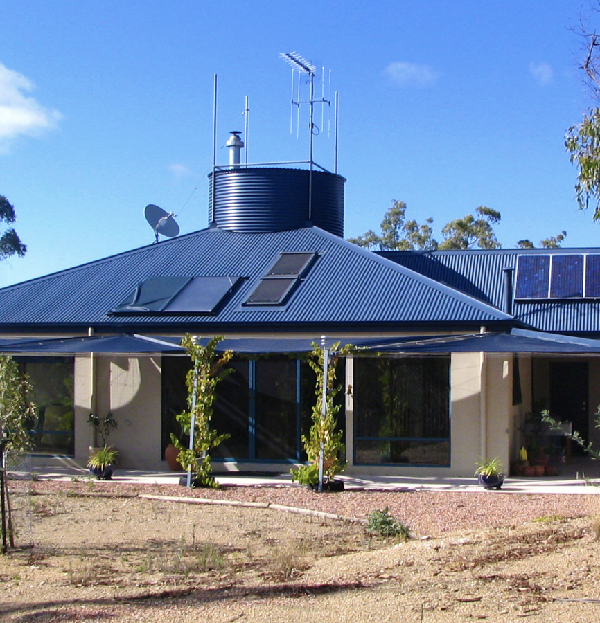 Modern house with a metal roof featuring solar panels and multiple antennas on top, with an outdoor patio area and dry, landscaped yard.