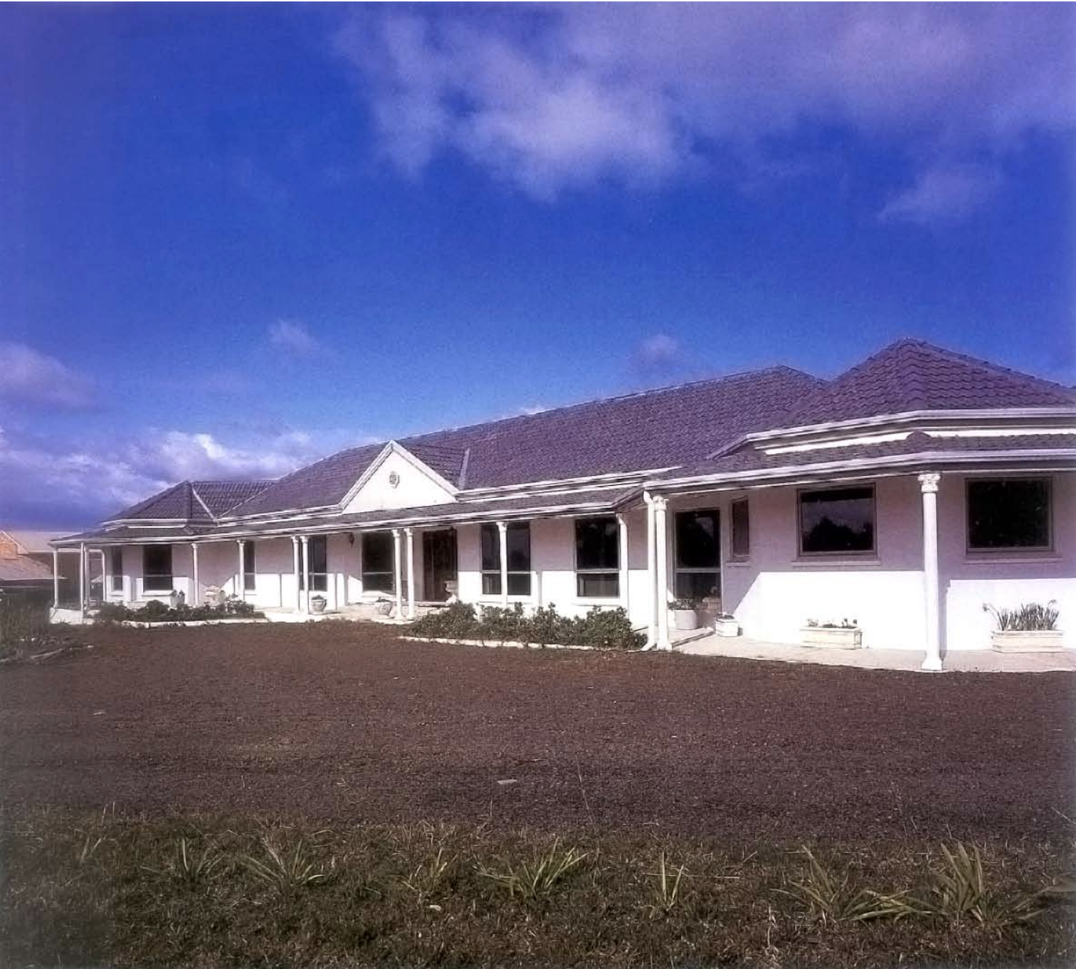 A white house with a porch and columns, multiple windows, a tiled roof, and a garden in front, against a background of a blue sky with clouds.