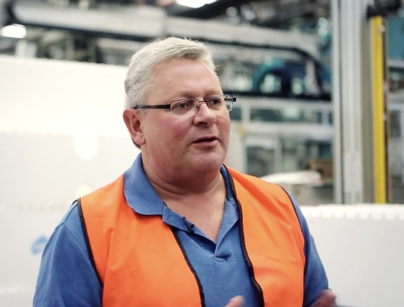 A middle-aged man wearing glasses, a blue polo shirt, and an orange safety vest in a manufacturing or warehouse setting.
