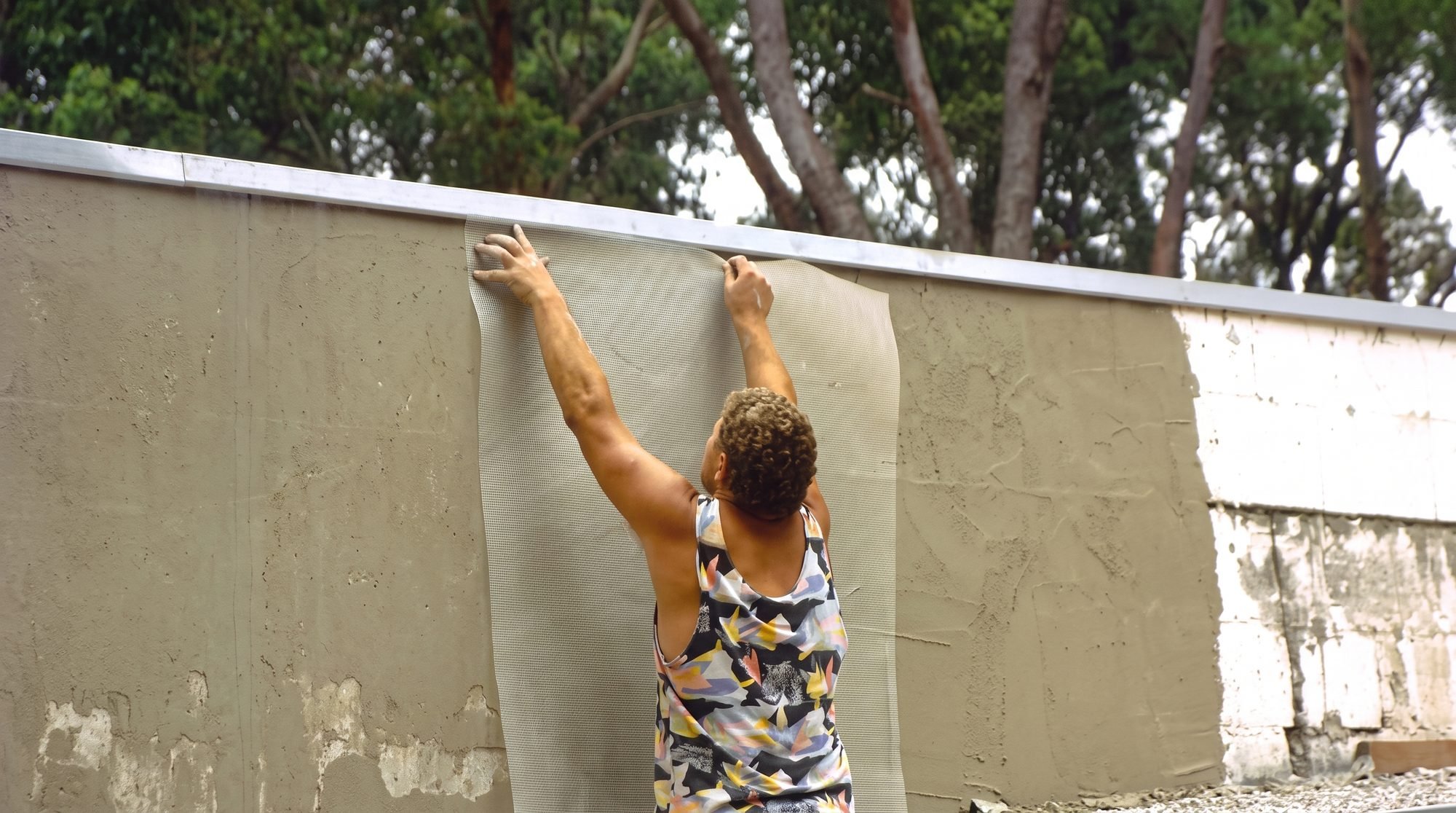A woman with curly hair attaching a screen or mesh to a beige concrete wall outdoors, with a background of trees and cloudy sky.