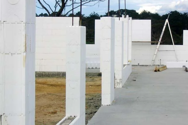 Construction site with white concrete block walls and a ladder, with trees and a cloudy sky in the background.