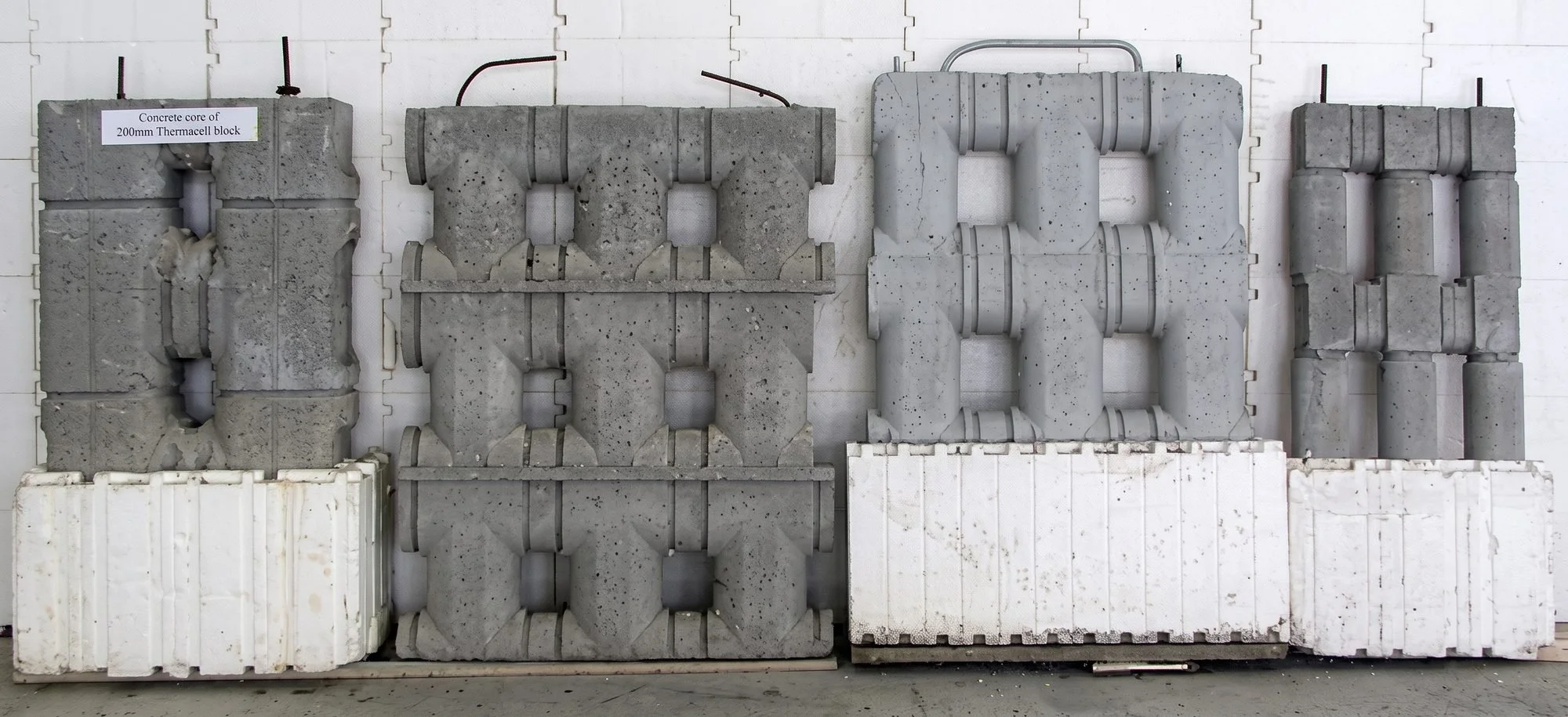 Various concrete blocks and molds stacked against a white wall, with a sign indicating one block is a 200mm Thermacell core.