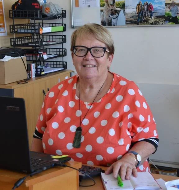 A smiling woman with glasses and short blond hair sitting at a desk with a laptop, notebook, and a pen. She is wearing a bright red polka dot shirt and a green pendant necklace. Behind her are shelves with office supplies and a wall with photos.