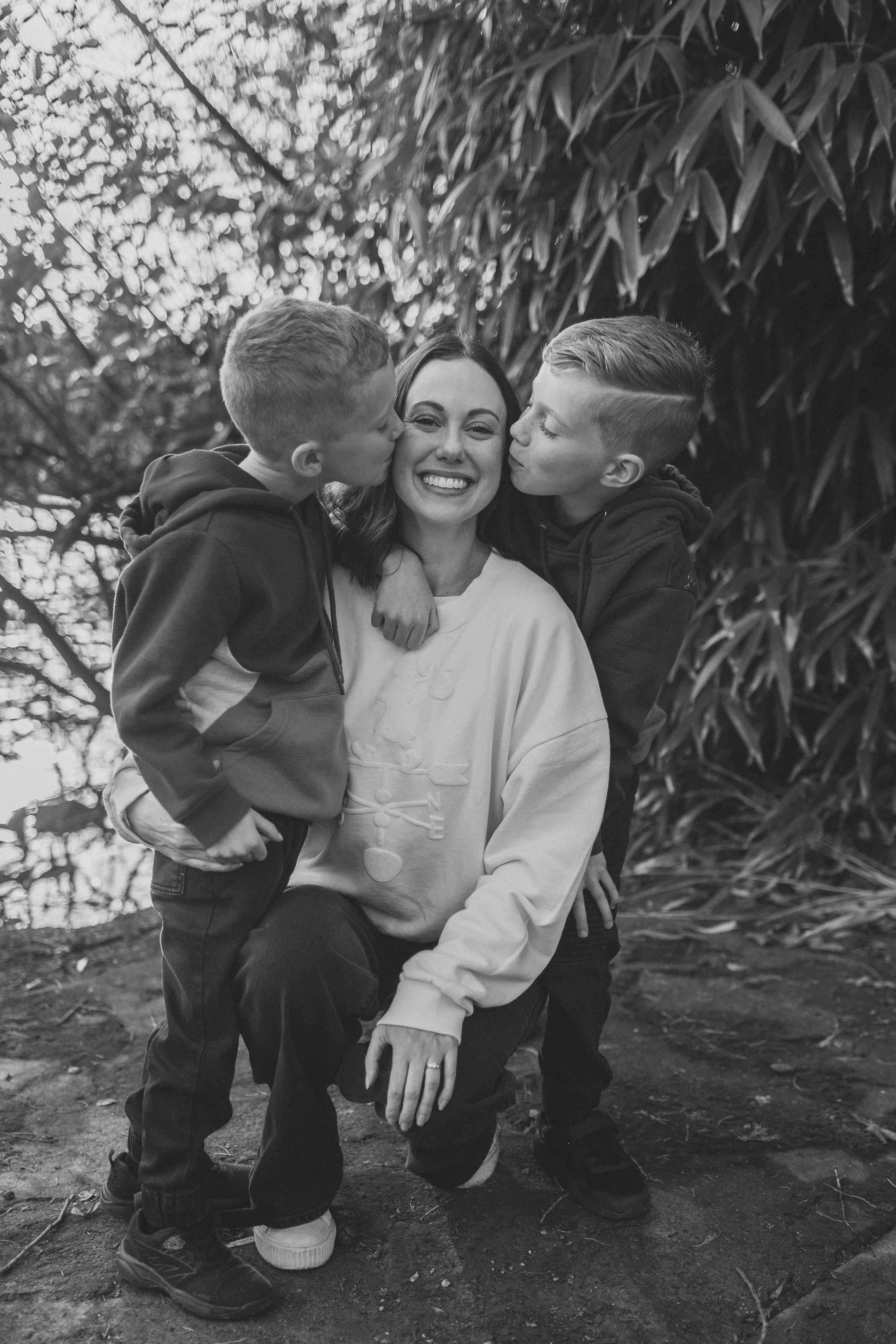 A woman with two young boys, one on each side, in an outdoor natural setting. The boys are giving her kisses on her cheeks, and she is smiling happily. The background has trees and foliage.