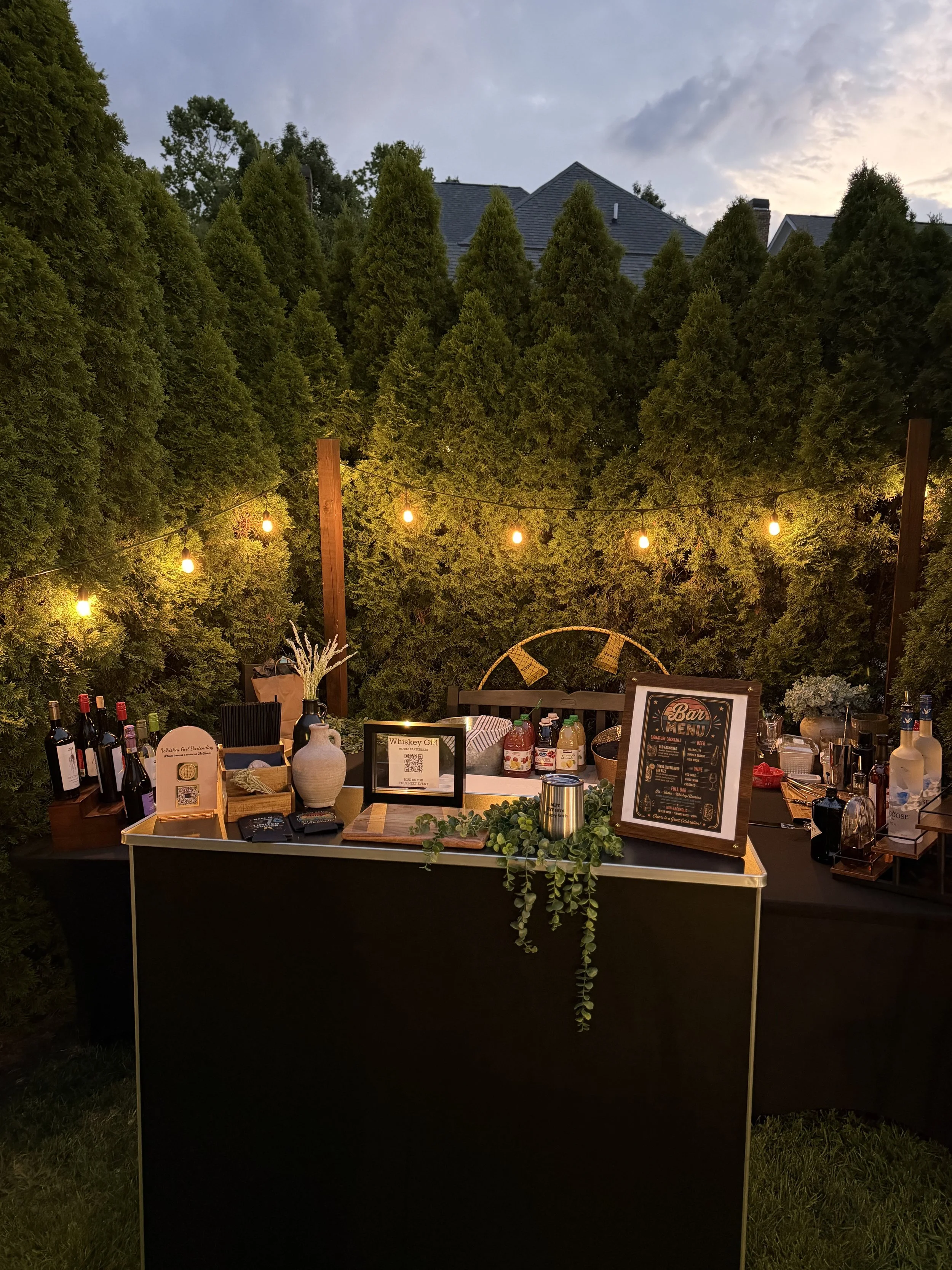 Outdoor bar setup with bottles of wine, alcohol, and mixers, illuminated string lights, greenery, and a sign showcasing the bar menu, set against a backdrop of tall trees and a cloudy sky at dusk.
