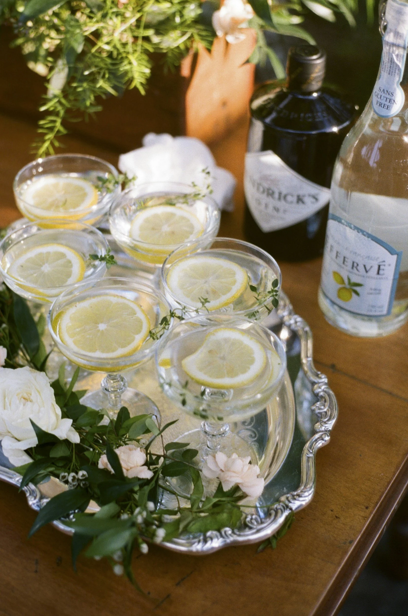 Elegant silver tray with multiple glasses of lemon-infused water, decorated with small sprigs of herbs, surrounded by white flowers and greenery on a wooden table.