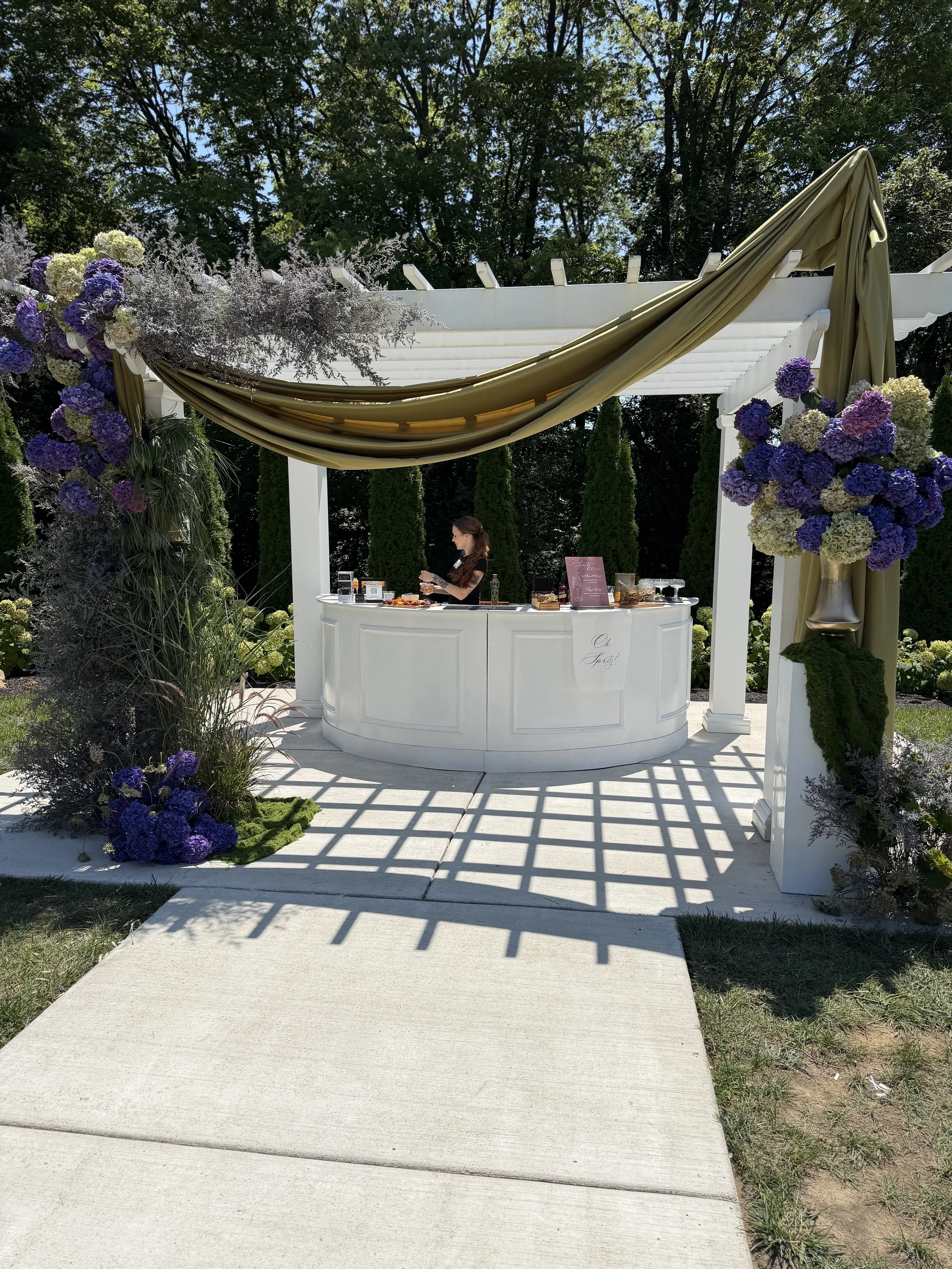 Outdoor wedding altar decorated with purple, white, and cream hydrangeas, set under a pergola with draped fabric, surrounded by greenery and trees.