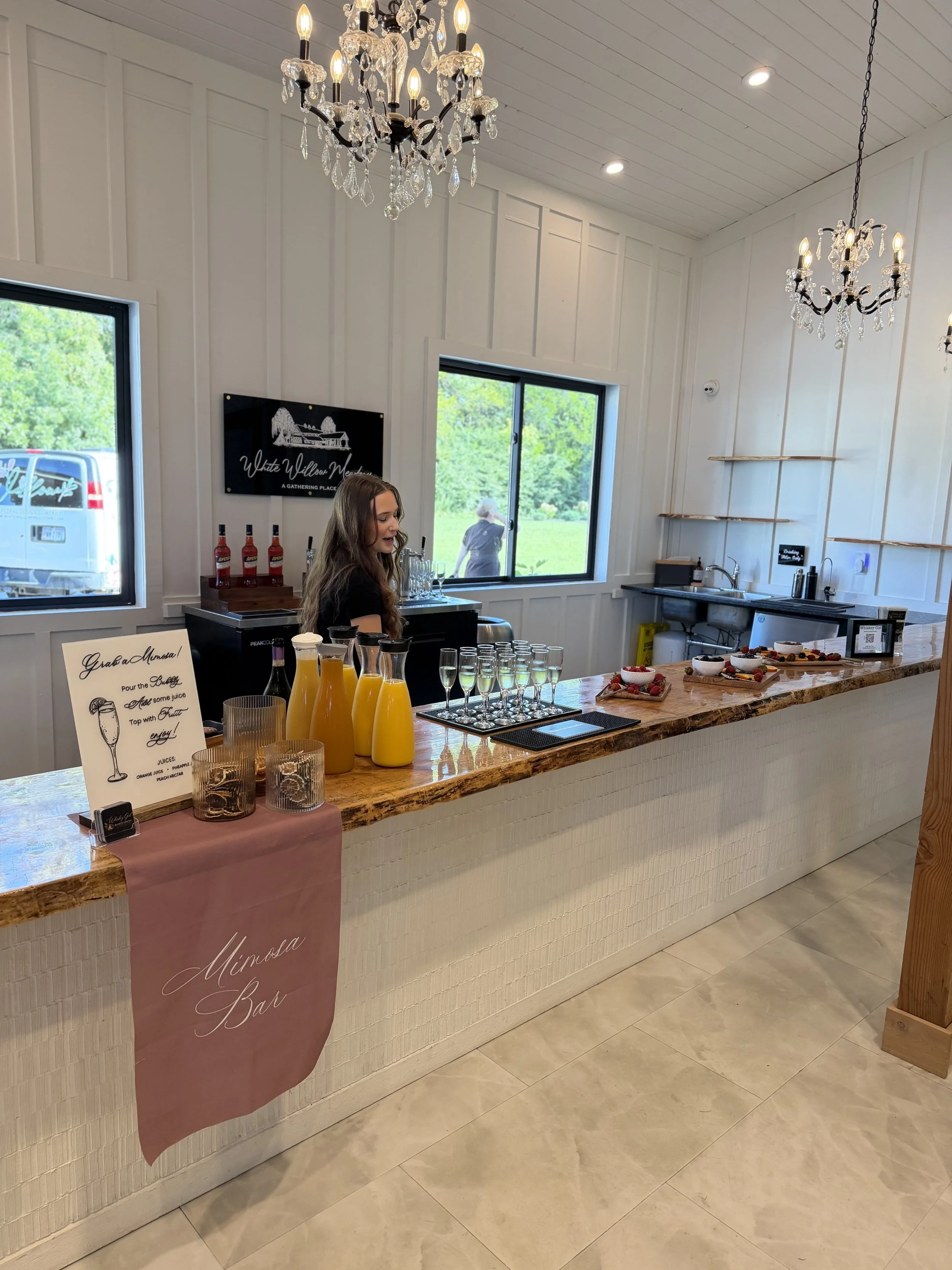 A reception area at a venue with a bar counter, hanging chandeliers, windows looking out onto greenery, and a sign reading 'White Willow Meadow A Gathering Place'.