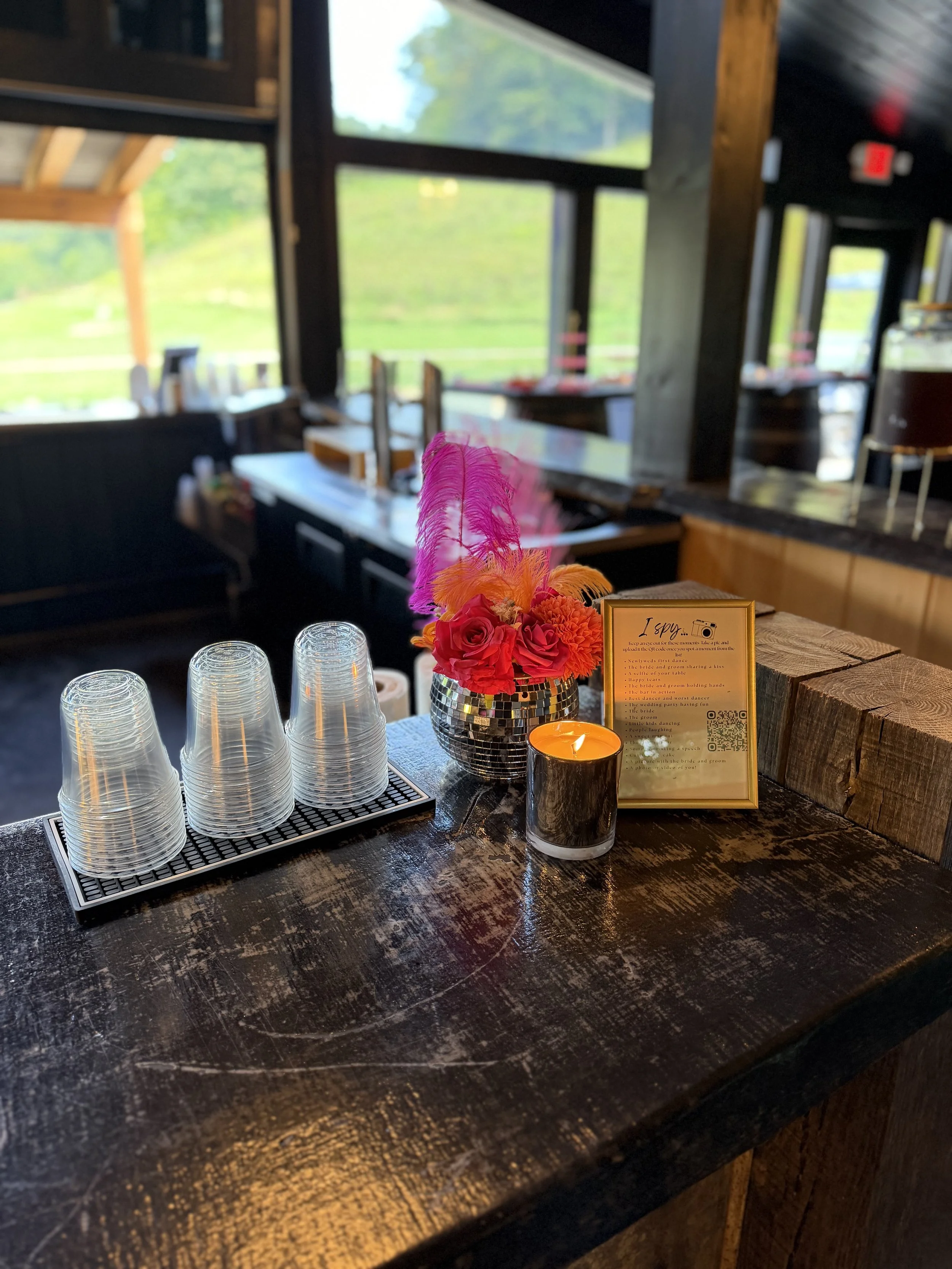 Close-up of a restaurant or bar counter with stacked clear glasses, a small flower arrangement with pink and orange feathers, a lit candle in a black holder, and a menu card on a wooden block. The background shows large windows with greenery outside.