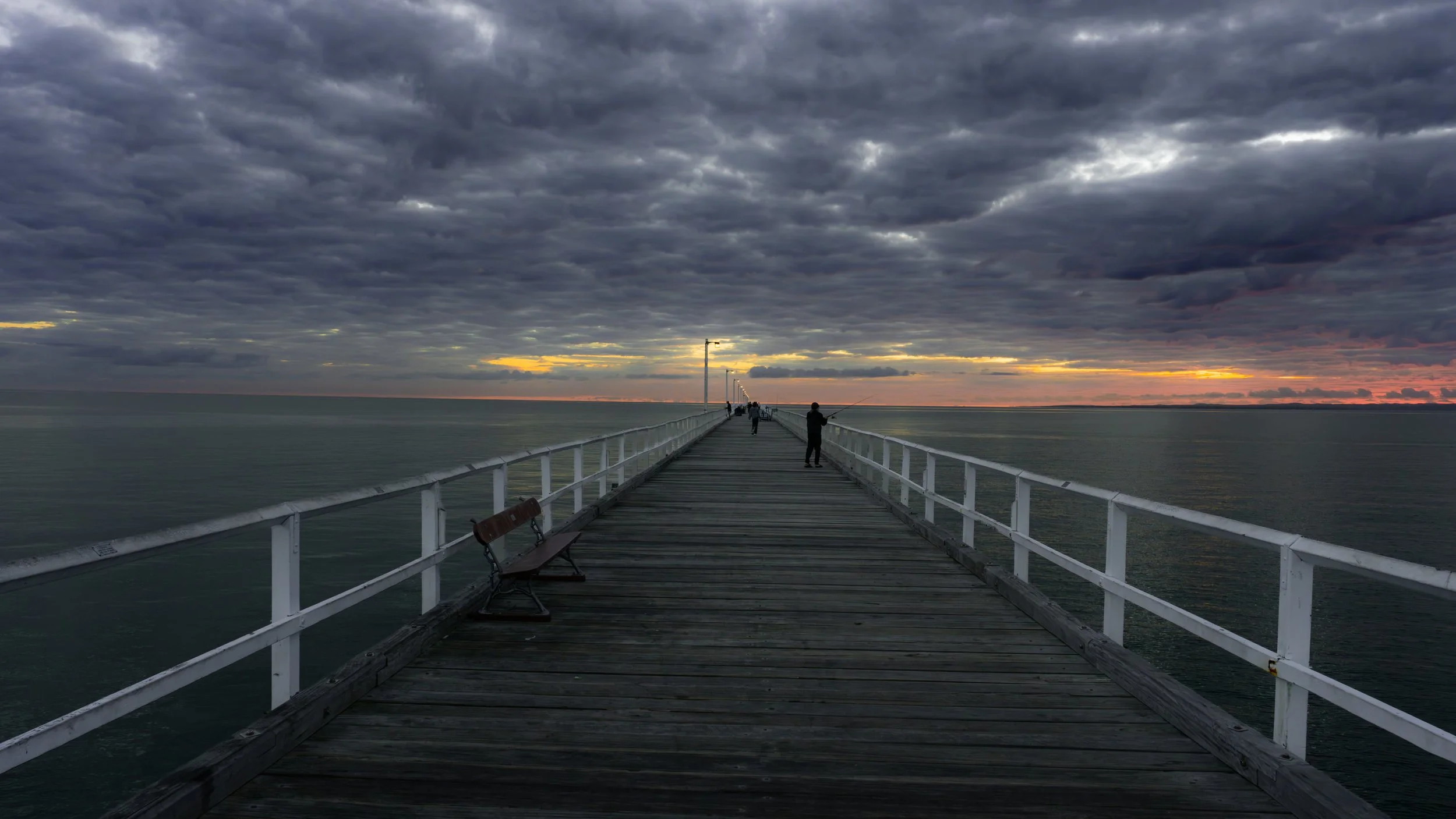 Wooden pier extending beneath dramatic storm clouds and a narrow band of sunset light.