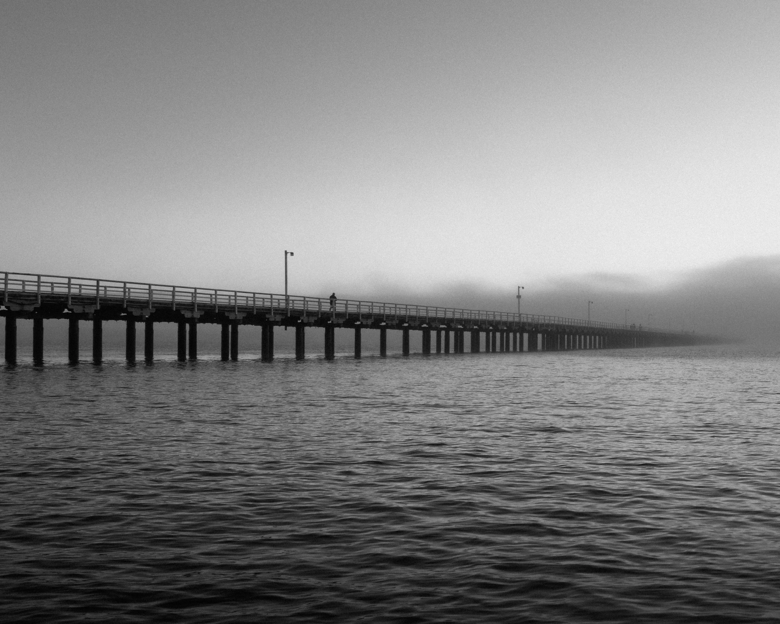 A solitary figure on a long pier disappearing into dense coastal fog, photographed in black and white.