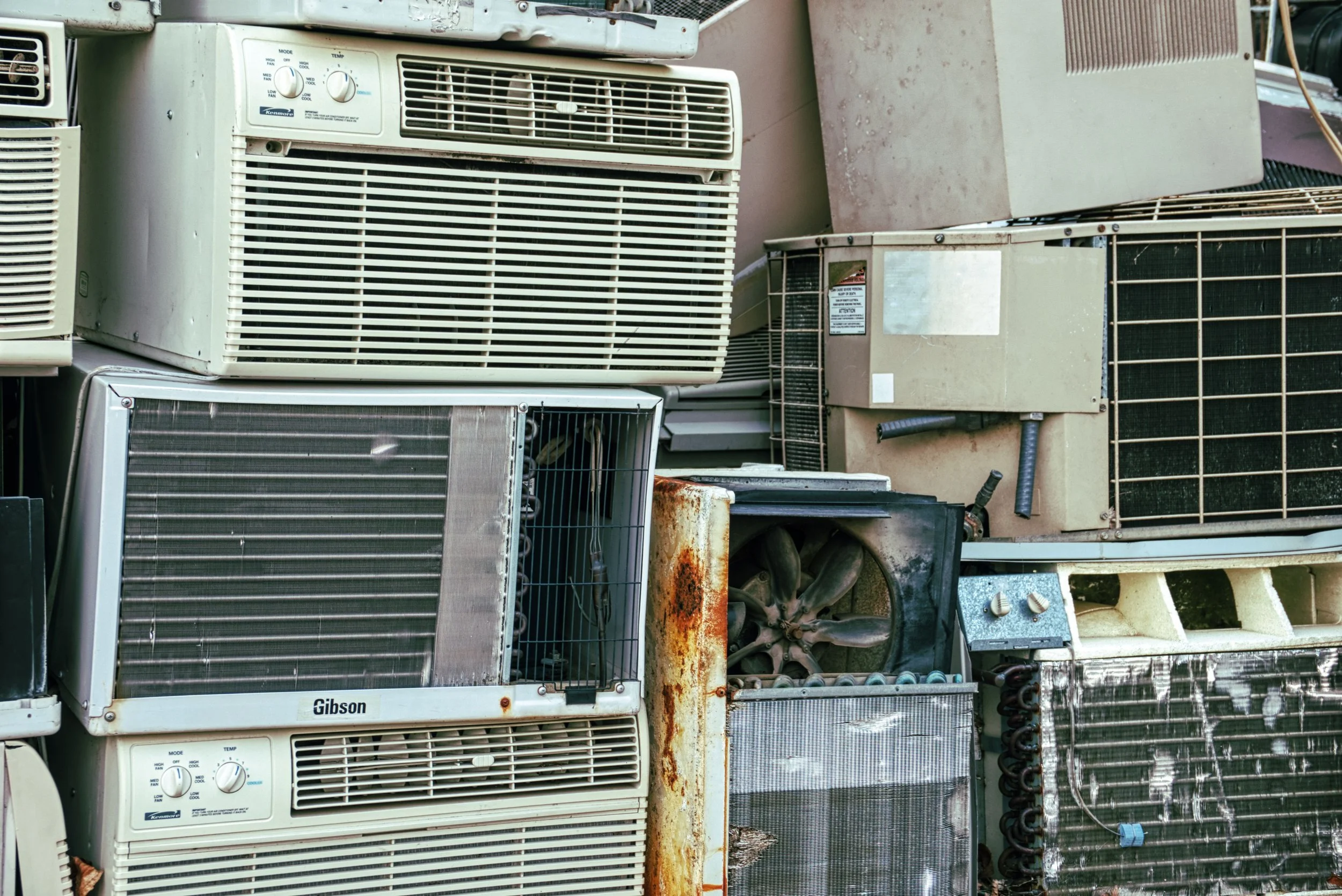 Four old white household appliances, including a refrigerator, stove, washing machine, and dryer, placed on the sidewalk in front of a suburban brick house with green bushes and trees.