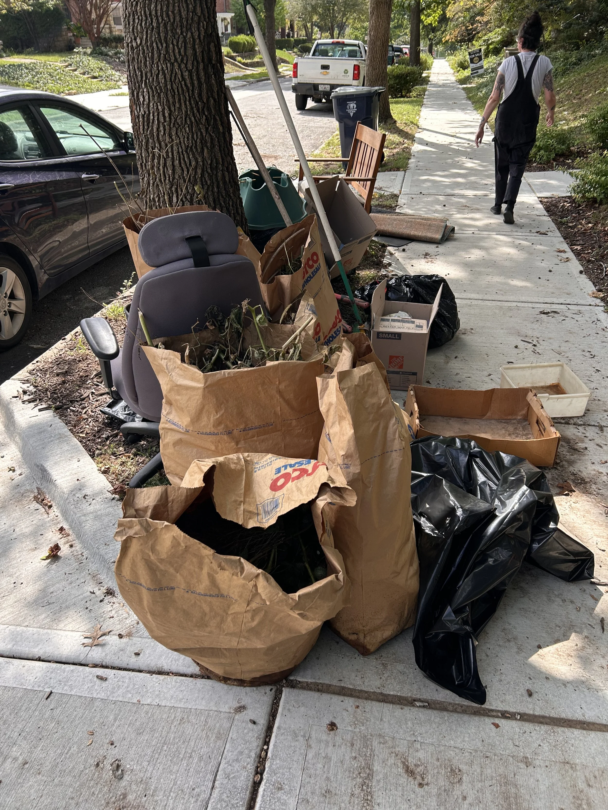 Leaves, branches, and firewood piled on the curb in front of a house with a brick exterior, surrounded by green bushes and trees.