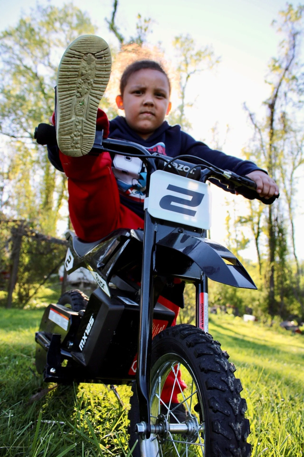 Child on a dirt bike with one foot raised, outdoors in a grassy area with trees, looking at the camera with a serious expression.