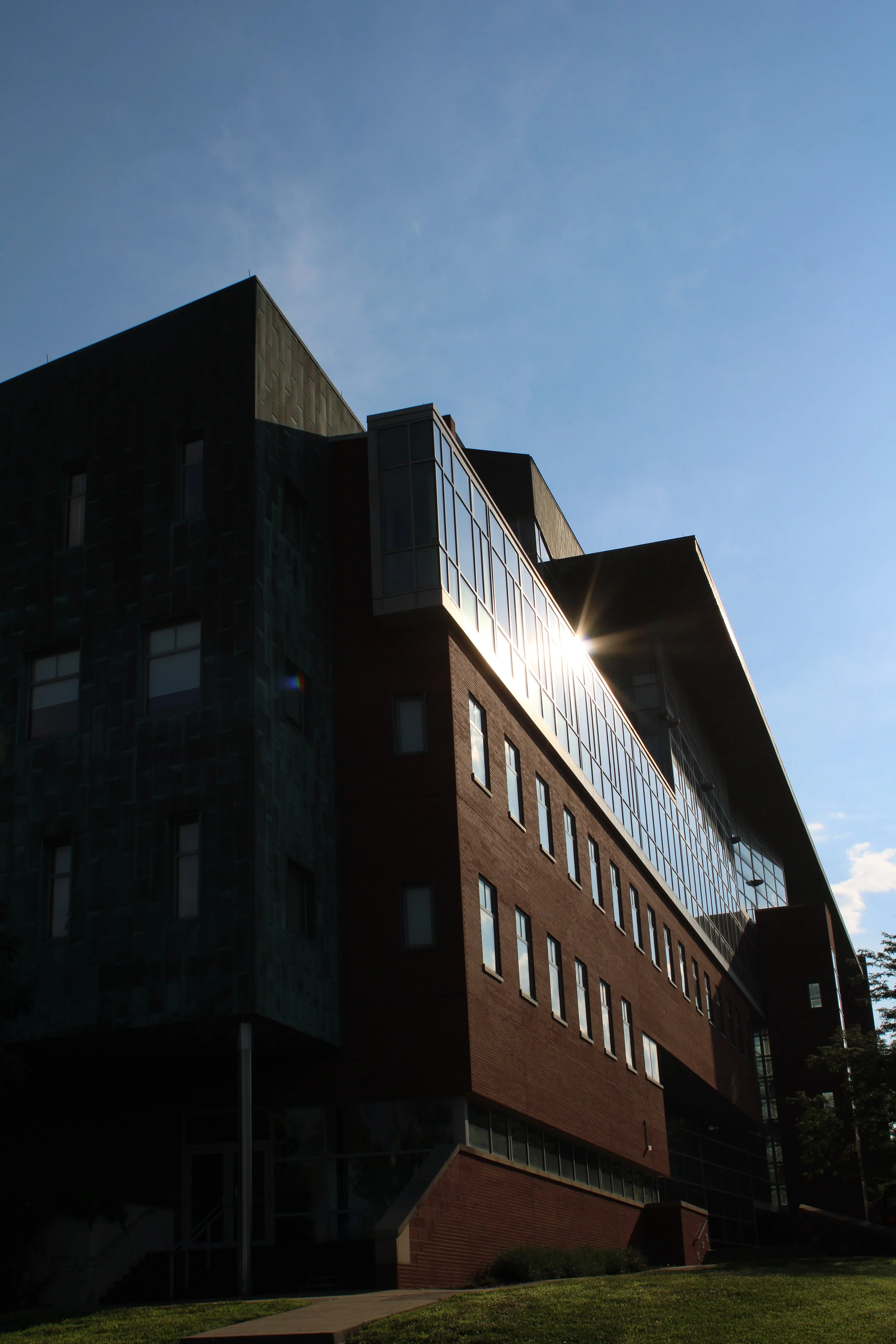 Low-angle view of a modern multi-story building with large windows and brick exterior against a clear blue sky with the sun shining brightly.