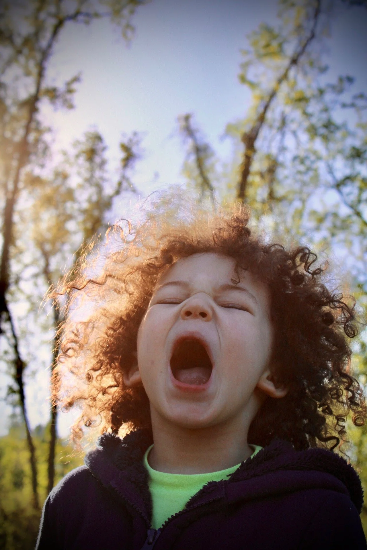 A young child with curly hair smiling and yelling outdoors with trees and sunlight in the background.