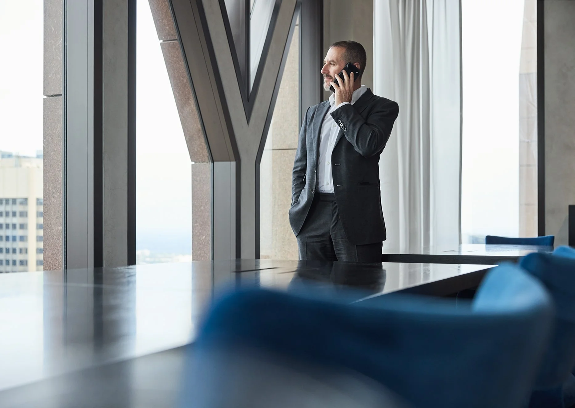 A man in a black suit with a white shirt, standing in an office near large windows, talking on a cell phone.