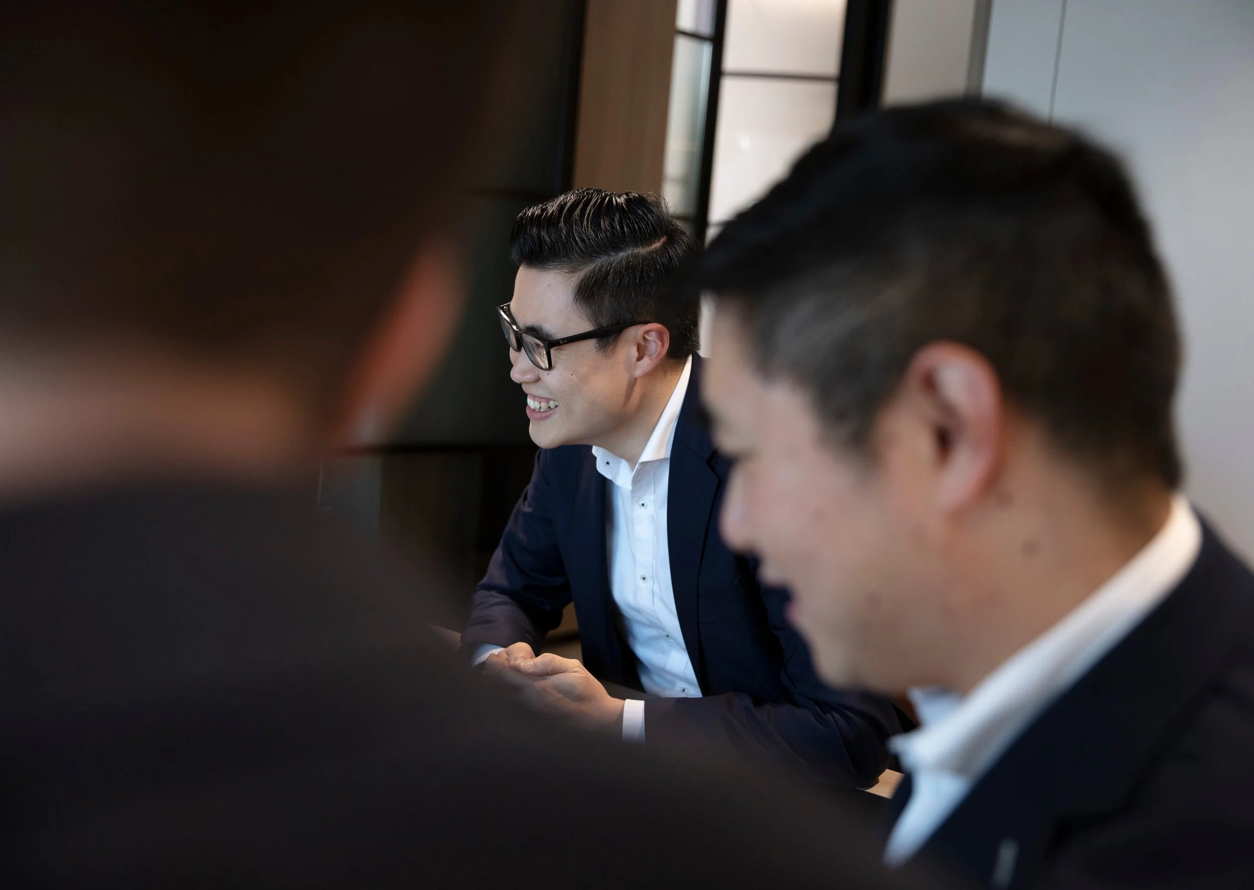 Smiling man with glasses in a business suit interacting with others at a professional meeting or conference.