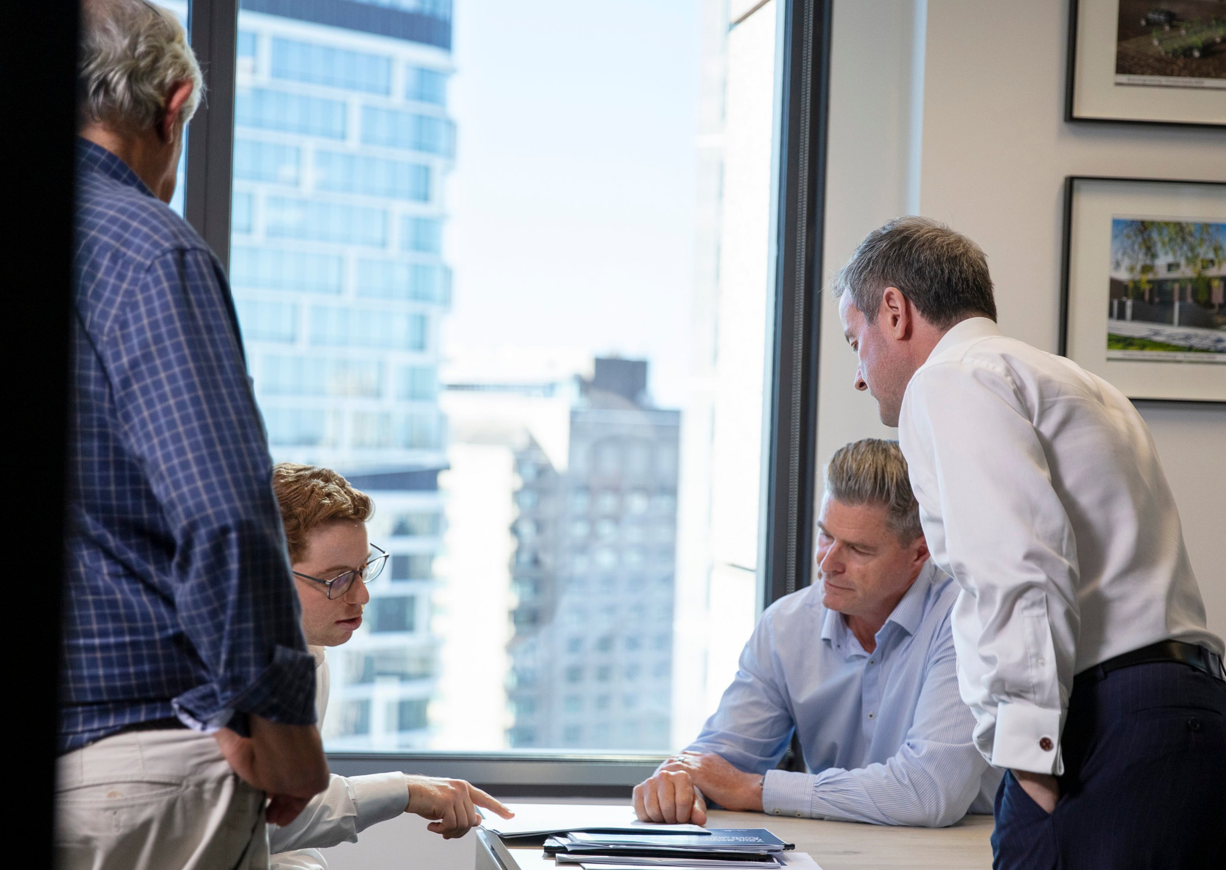 Group of professionals in a meeting, discussing around a table in a modern office with large windows and city buildings outside.