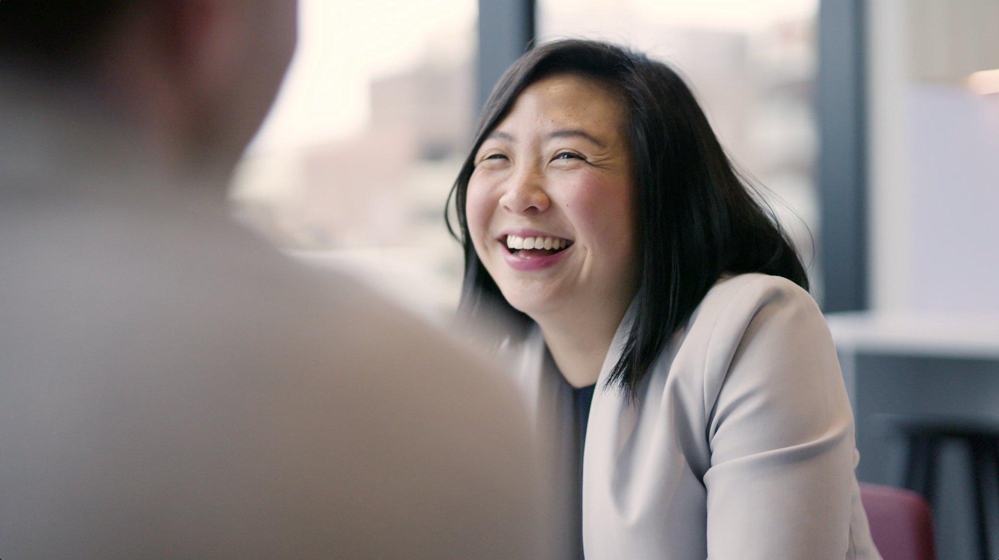 A woman with long black hair smiling and laughing during a conversation in an office setting.