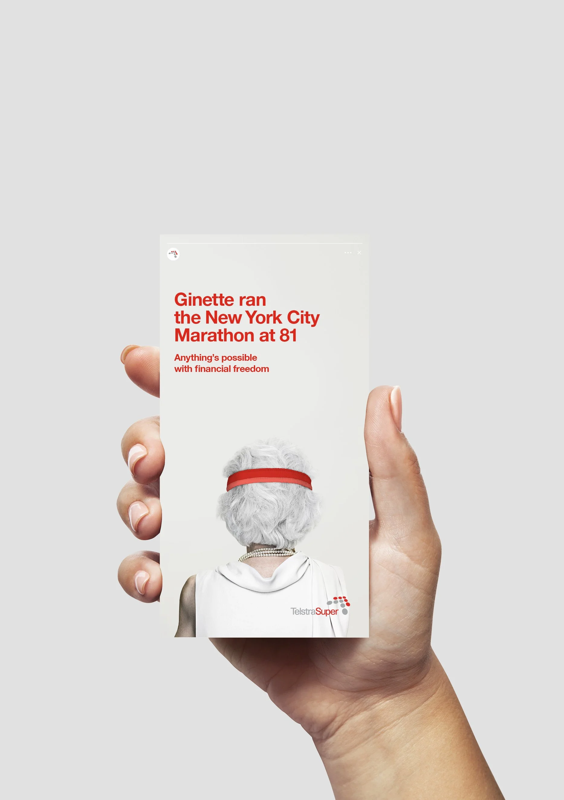 A person's hand holding a brochure or flyer with the back side showing. The brochure features a silver-haired elderly woman with a red headband, wearing pearl necklaces and a white top, with the text "Ginette ran the New York City Marathon at 81" and the phrase "Anything's possible with financial freedom." There is also a partial logo that reads "Telstra Super" in the lower right corner.