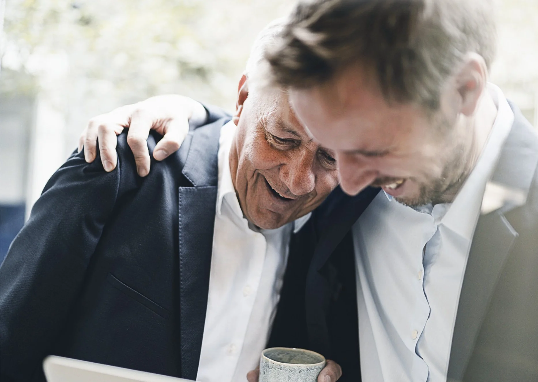 Two men sharing a moment of joy, one with his arm around the other, in an indoor setting with soft natural lighting.