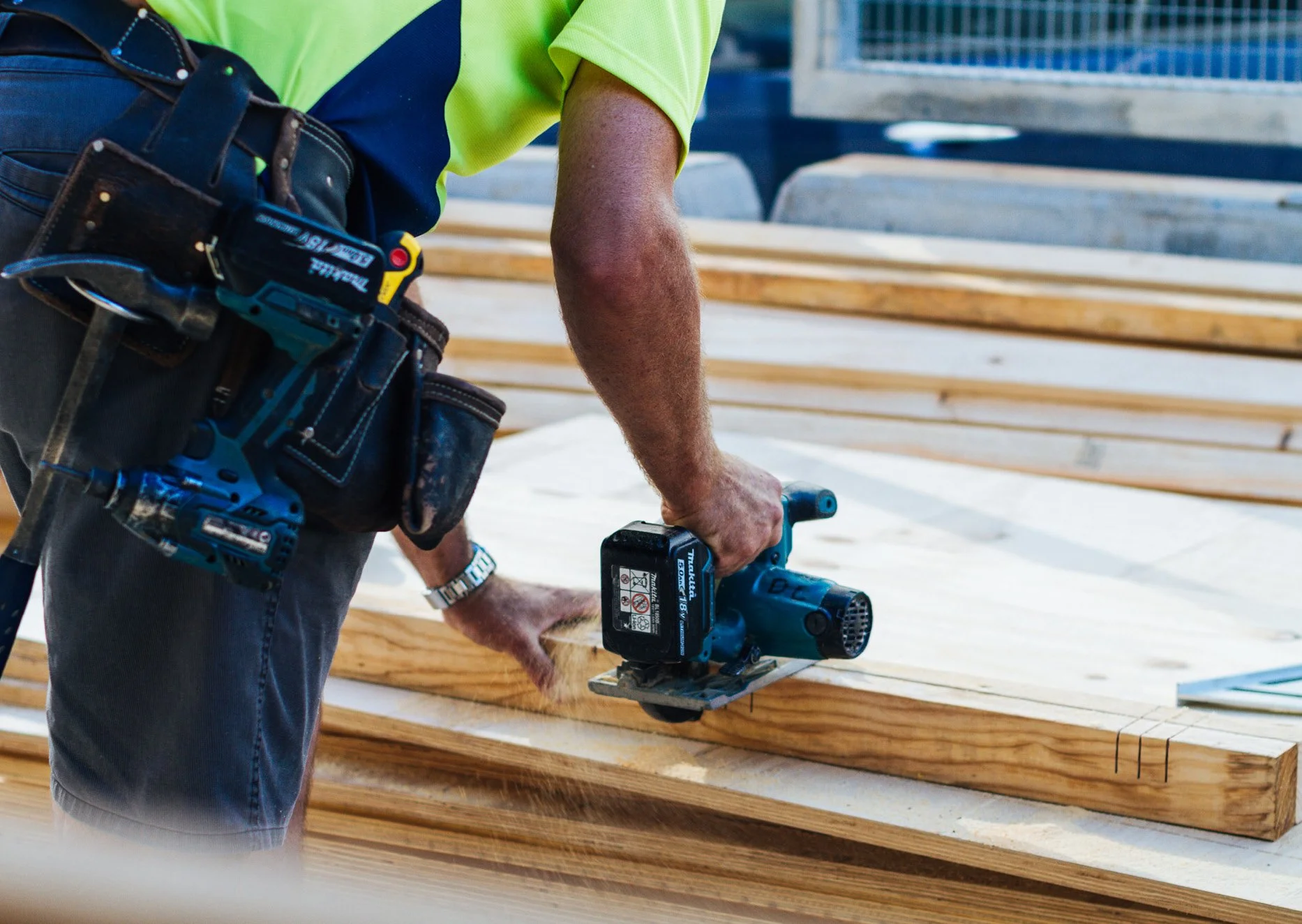 A construction worker using a power saw on wooden planks at a construction site, wearing a bright yellow safety shirt and tool belt.