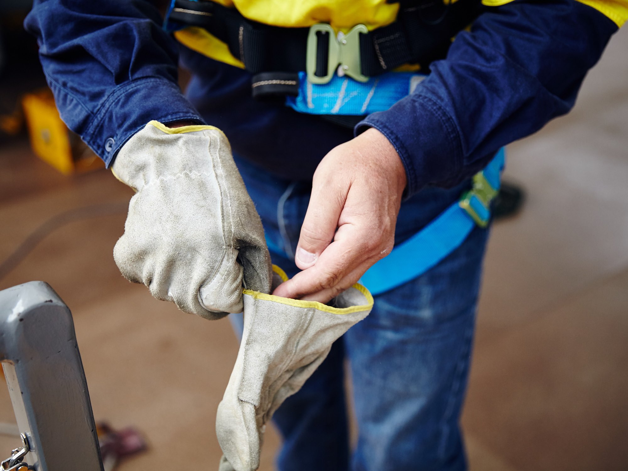 Person wearing blue work shirt and safety harness, holding and inspecting a pair of gloves.