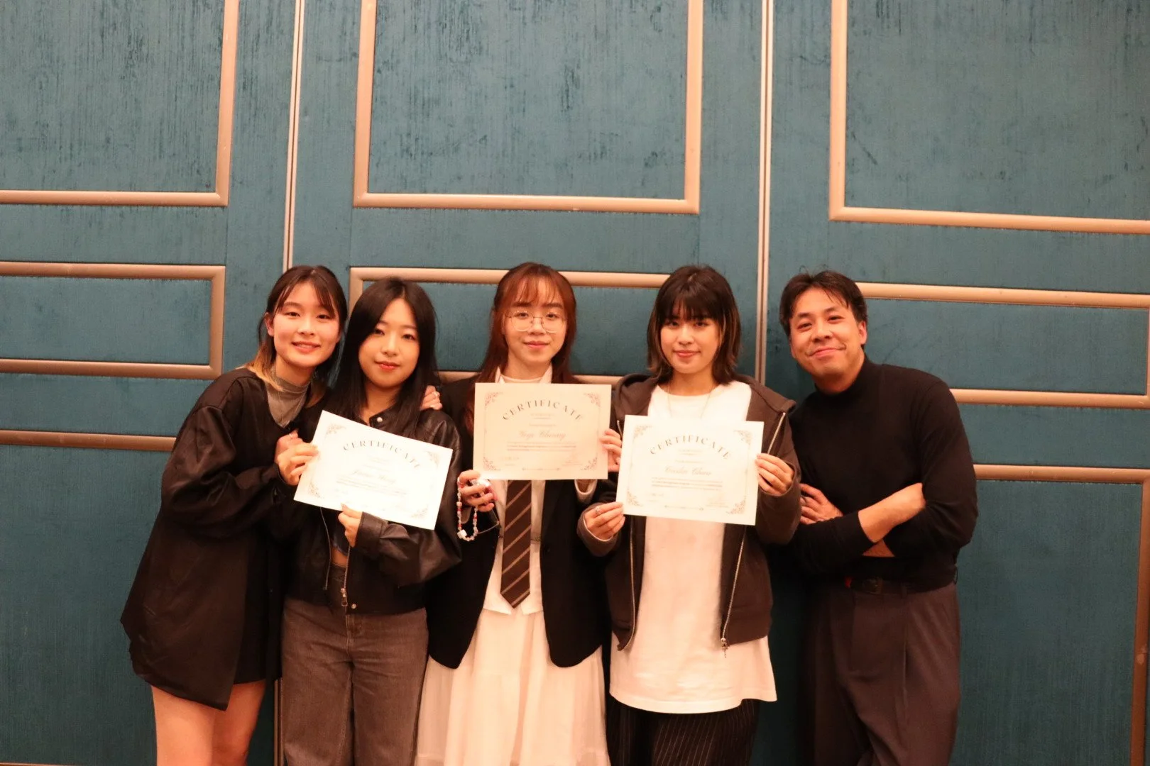 Group of five people, four women and one man, standing together in front of a teal wall. The women are holding certificates, smiling, and the man is standing on the far right with a smile.