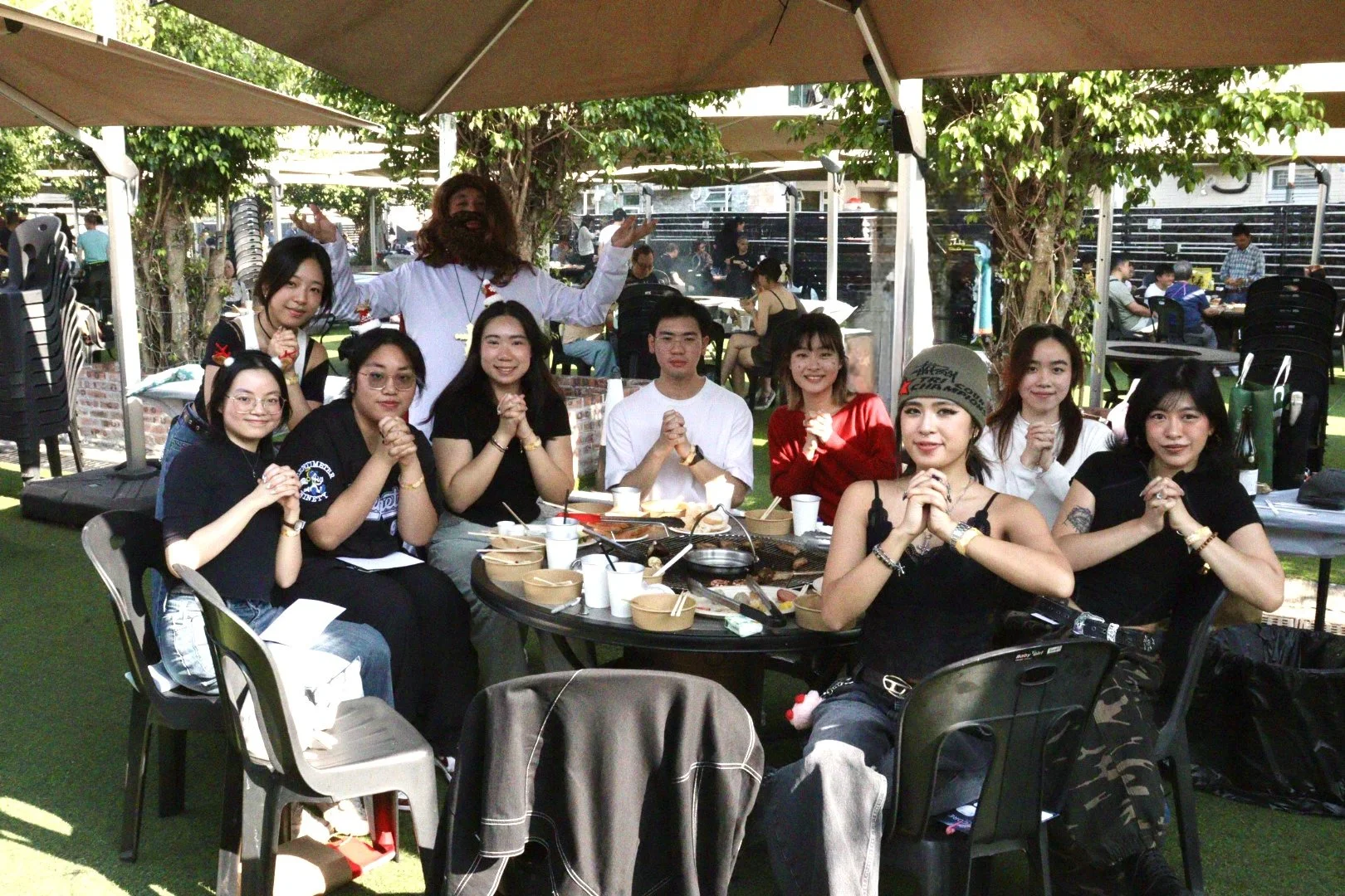 A group of people gathered around a table outdoors, enjoying a meal, with trees and other diners in the background.