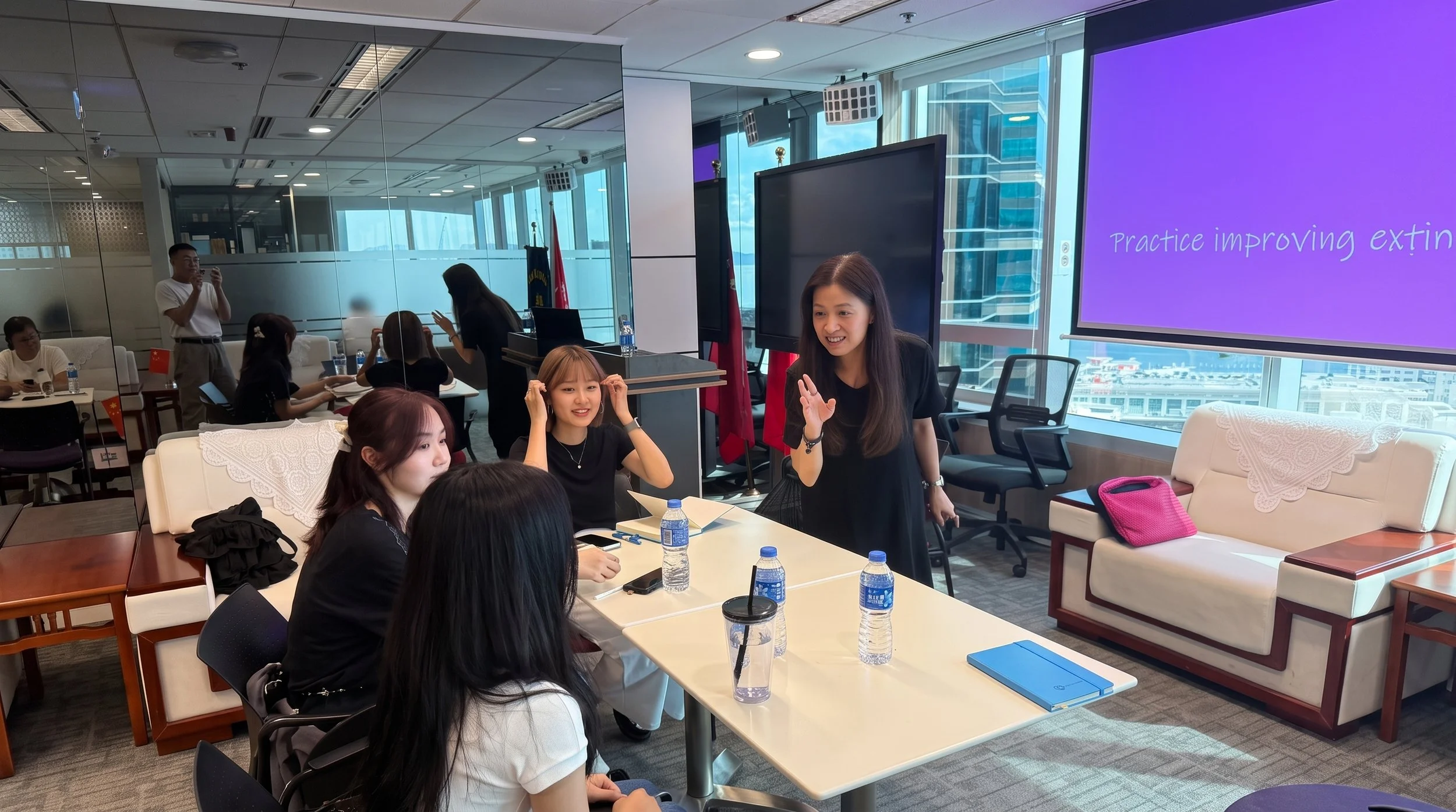 A group of people attending a workshop or meeting in a modern conference room with floor-to-ceiling windows, on which a woman is presenting or speaking, with a large purple screen displaying practice tips.