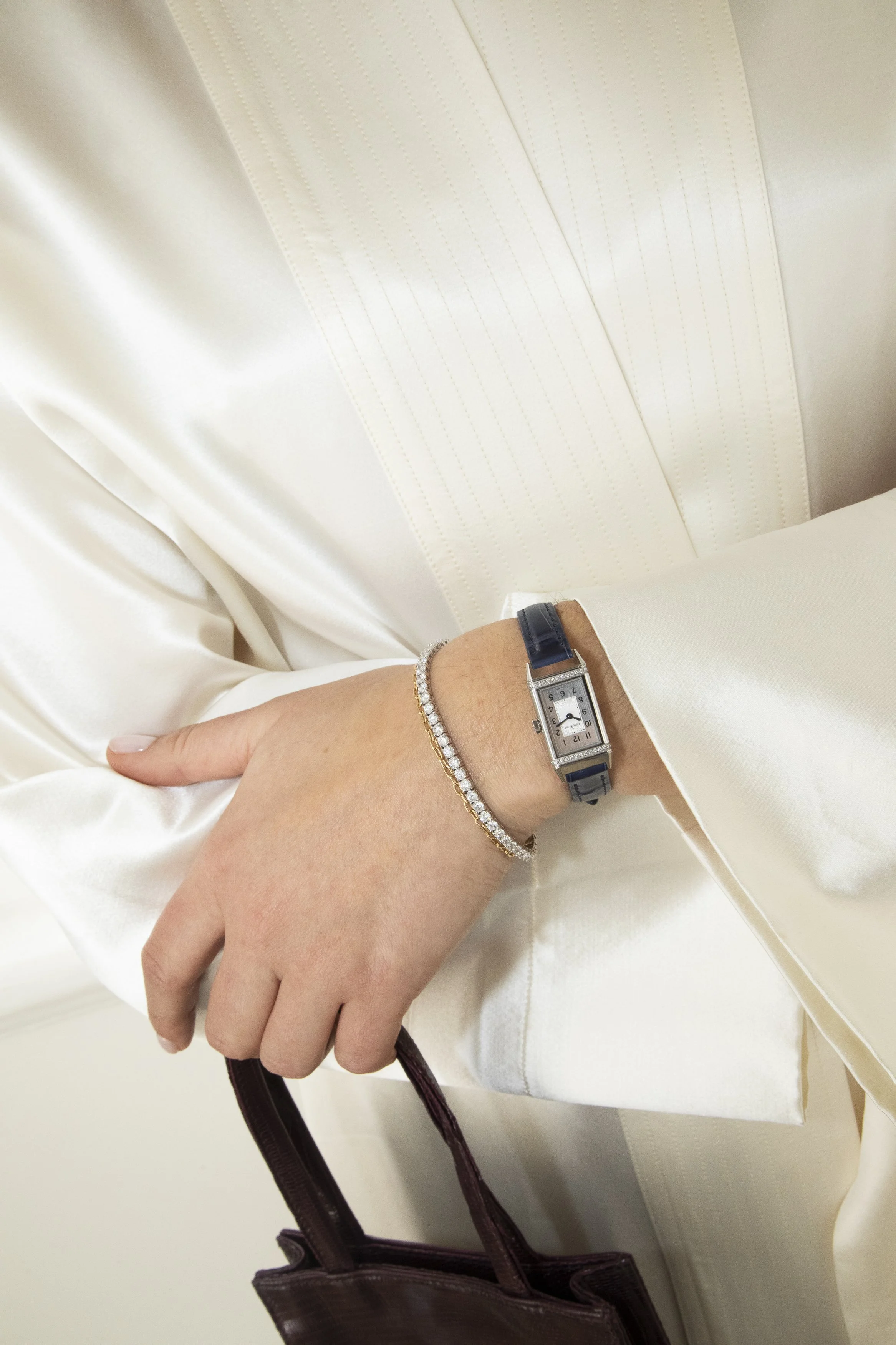 A person wearing a white satin blouse, a silver and blue wristwatch, and a diamond bracelet, holding a dark brown handbag with purple handles. The person's left hand is visible, with the thumb resting on the fingers and a beige-colored wall or surface in the background.