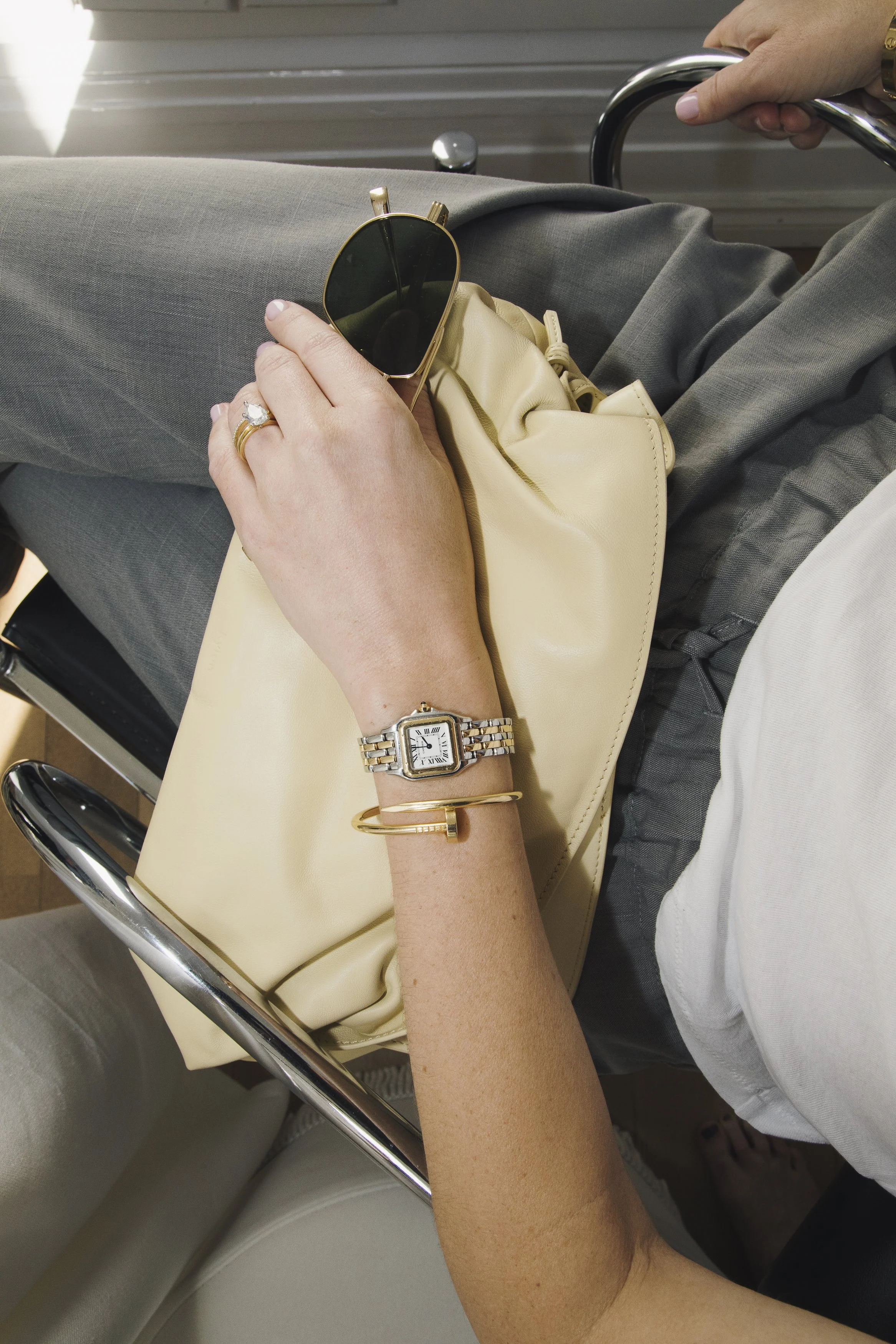 A person sitting in an airport or waiting area holds a pair of sunglasses and a beige purse on their lap. They wear a gold watch, a gold bracelet, a ring, and a wristwatch. The person is dressed in gray pants and a white shirt. The seating is cream-colored with metallic armrests.