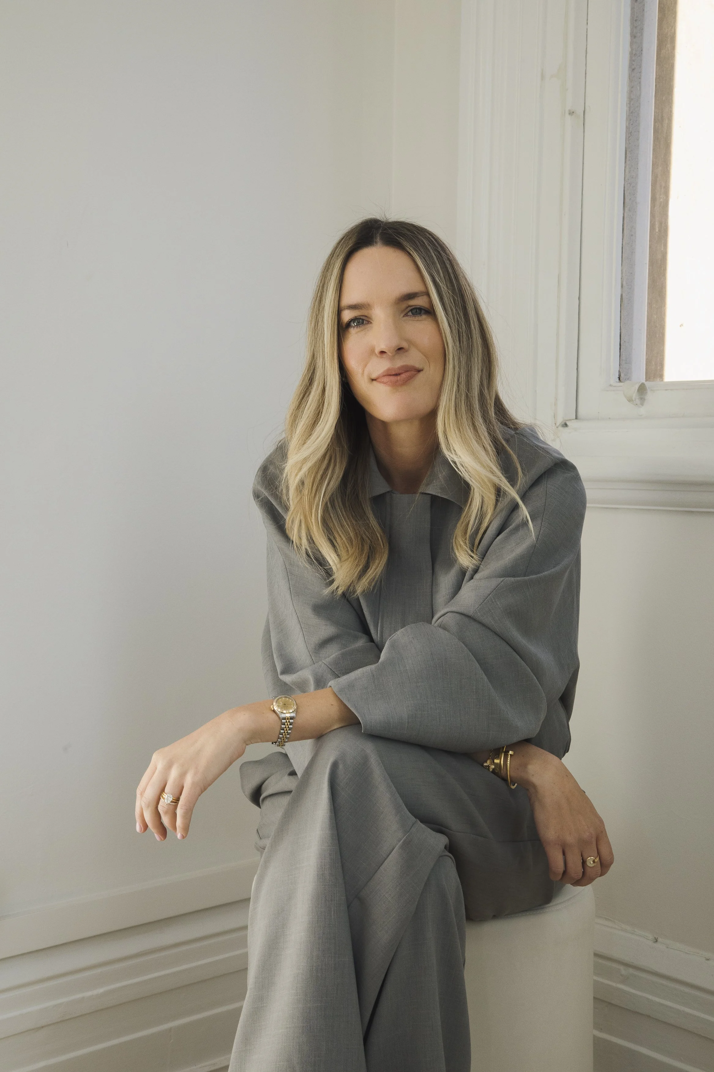 A woman with long blonde hair, wearing a grey suit, sitting in front of a white wall next to a window, with her arms crossed and smiling gently.