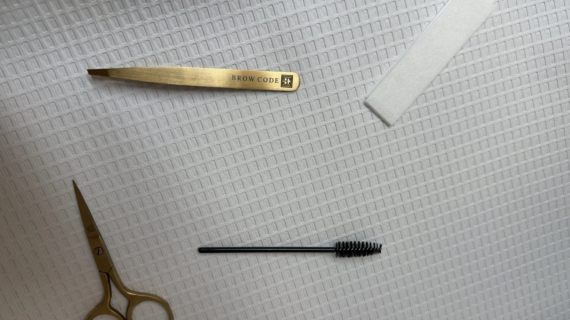 A pair of gold tweezers with the words 'BROW CODE' engraved on them, a small black spool of eyelash extension adhesive, a disposable eyelash brush, and a gold-colored scissors on a white textured surface.