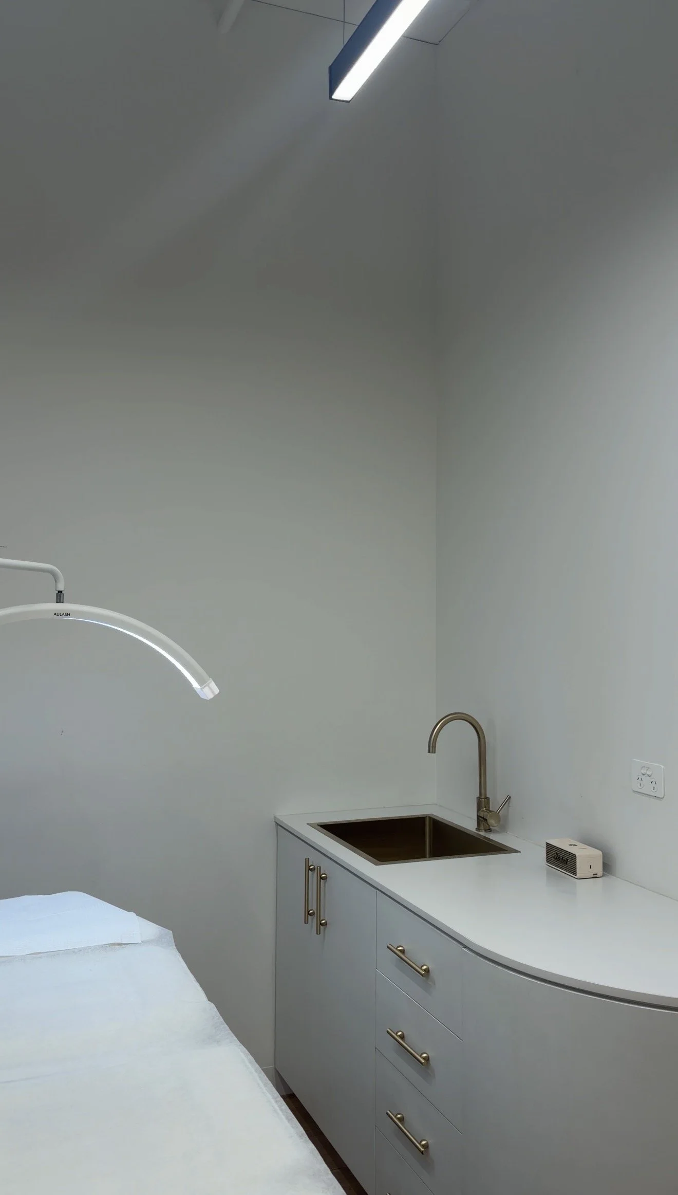 A clinical room with a white counter, a stainless steel sink with a gold faucet, white cabinets with gold handles, a wall-mounted lamp, and a medical device on the counter.