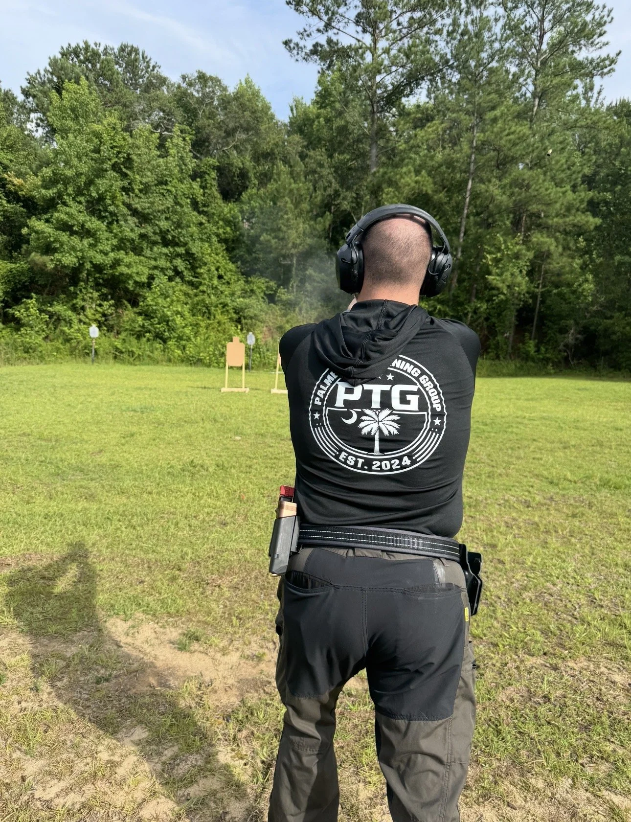 A person with a buzz cut hairstyle wearing black tactical clothing and ear protection aiming at targets during outdoor shooting practice.