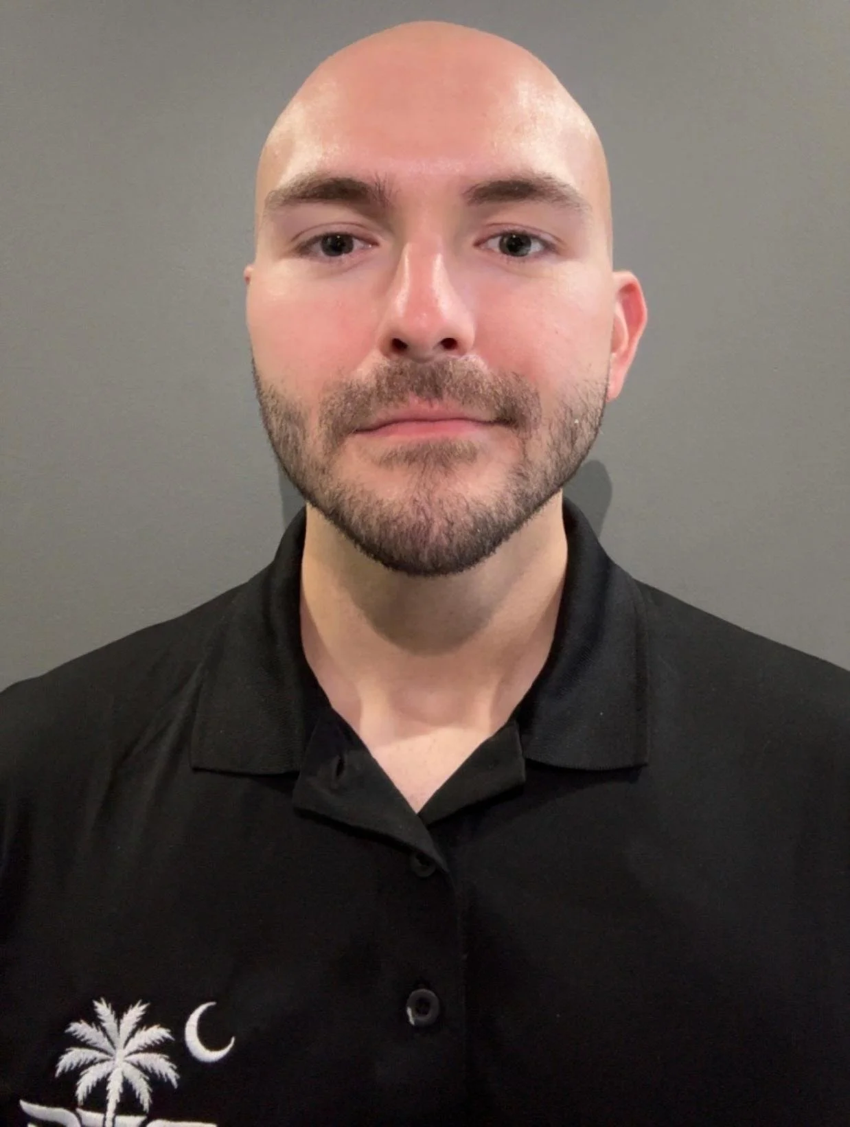 A bald man with a beard and mustache wearing a black polo shirt with a palm tree and moon logo, standing against a gray wall.