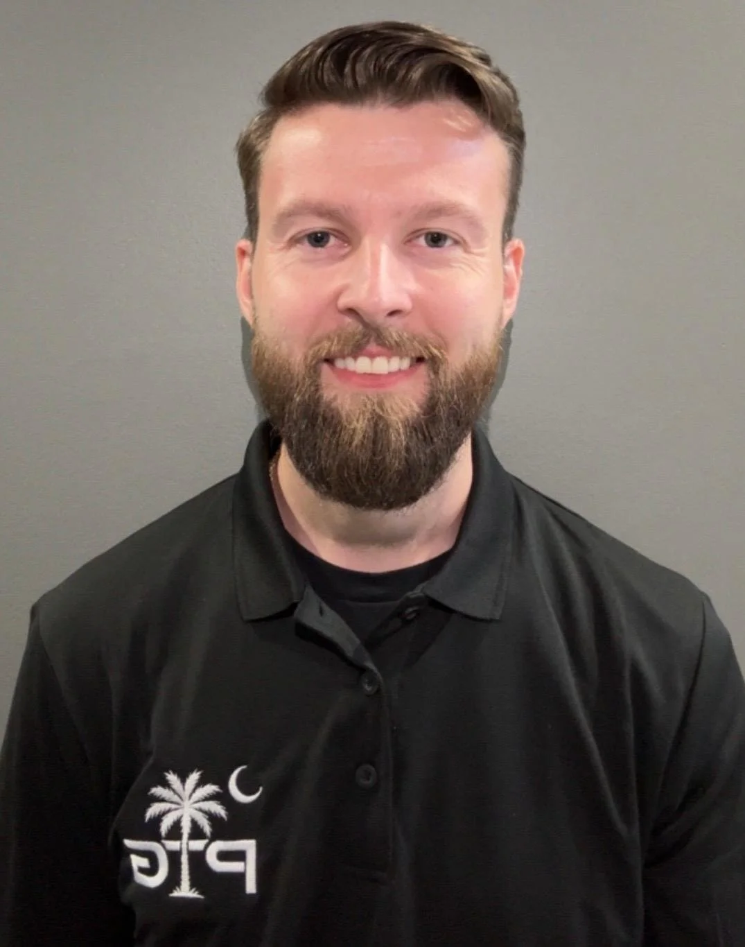 A smiling man with a beard and short brown hair wearing a black polo shirt with a palm tree logo on it.