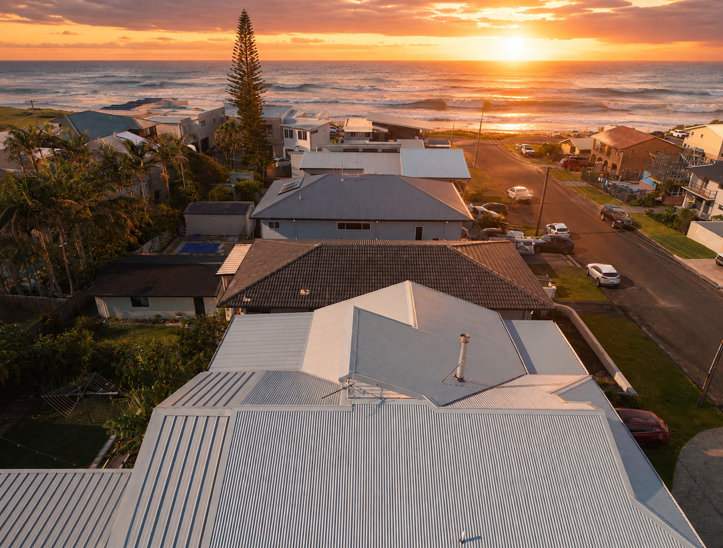 Aerial view of a coastal neighborhood at sunset with houses, palm trees, and the ocean in the background.