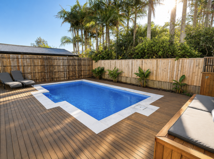Backyard with a small rectangular swimming pool, wooden deck, two lounge chairs, a wooden bench with cushions, and a bamboo fence surrounded by palm trees and other greenery.