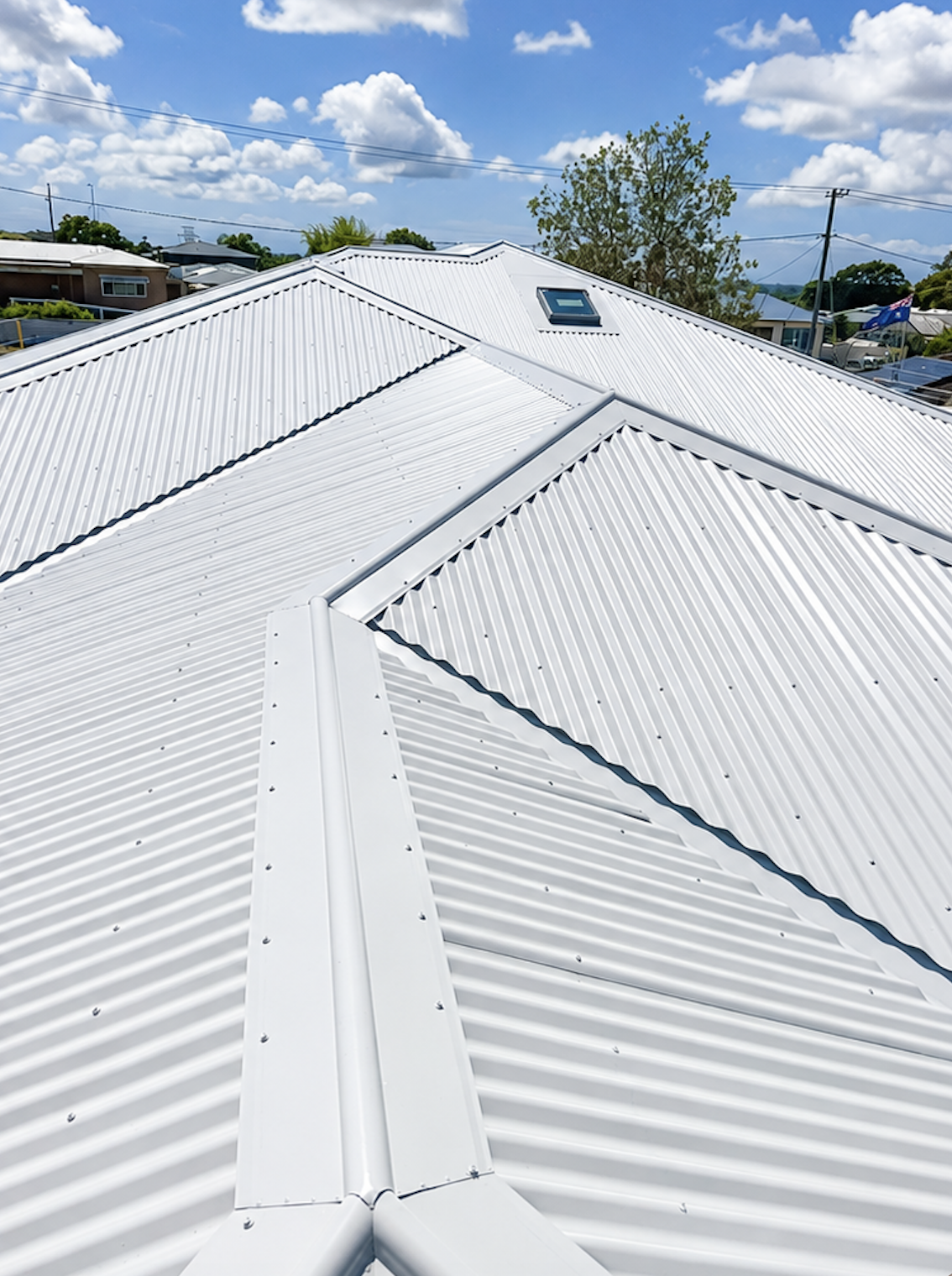View of a white metal roof with a skylight, under a blue sky with scattered clouds in a residential neighborhood.