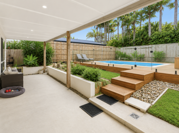 Backyard patio with a small swimming pool, wooden deck, lounge chairs, lush greenery, and a privacy fence with palm trees in the background.