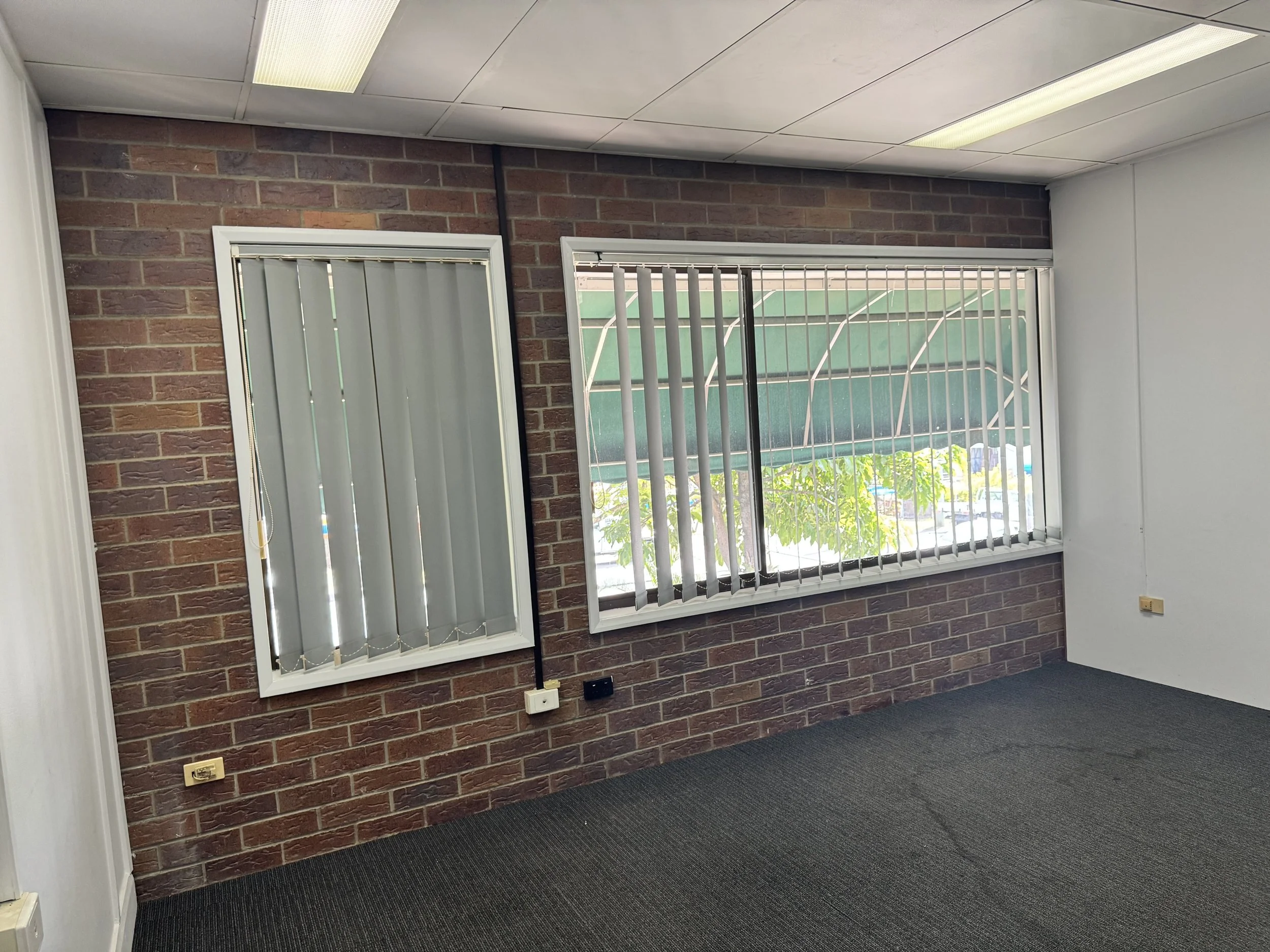 An office interior with a brick wall, two windows with blinds, a gray carpet, and ceiling lights.