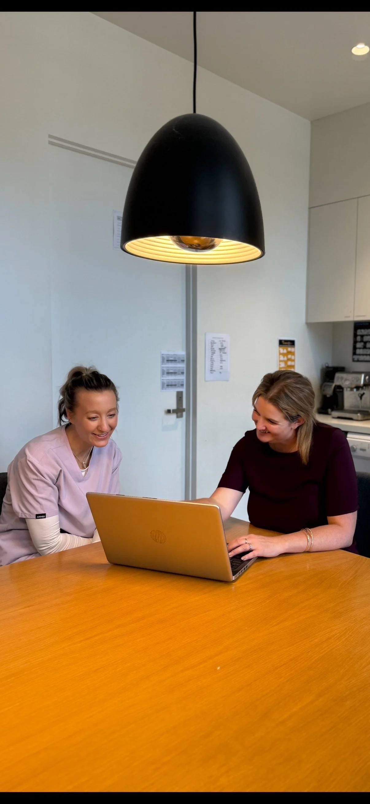 Two women sitting at a wooden table, smiling and looking at a yellow laptop in a room with white walls and a large black pendant light hanging above.