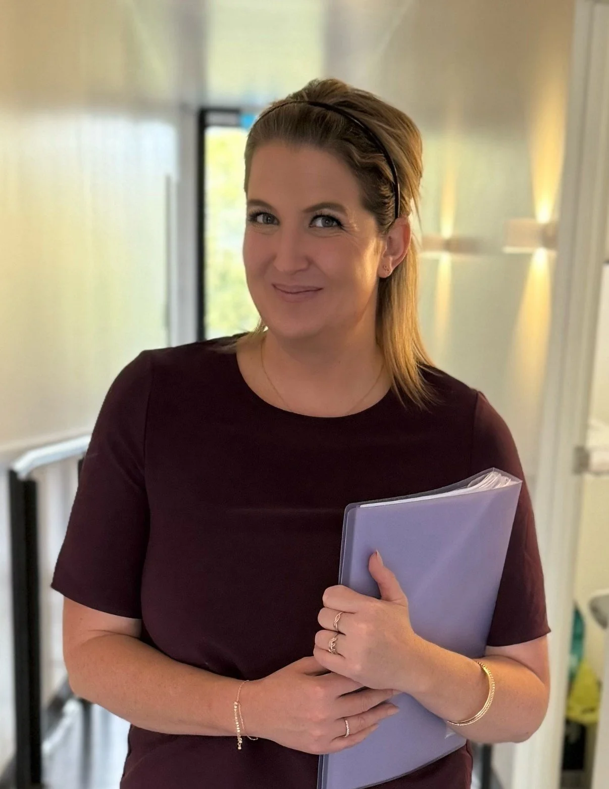 A woman with light brown hair and a black headband, wearing a dark red dress, holding a purple notebook, standing in a brightly lit indoor space with yellow and white walls.