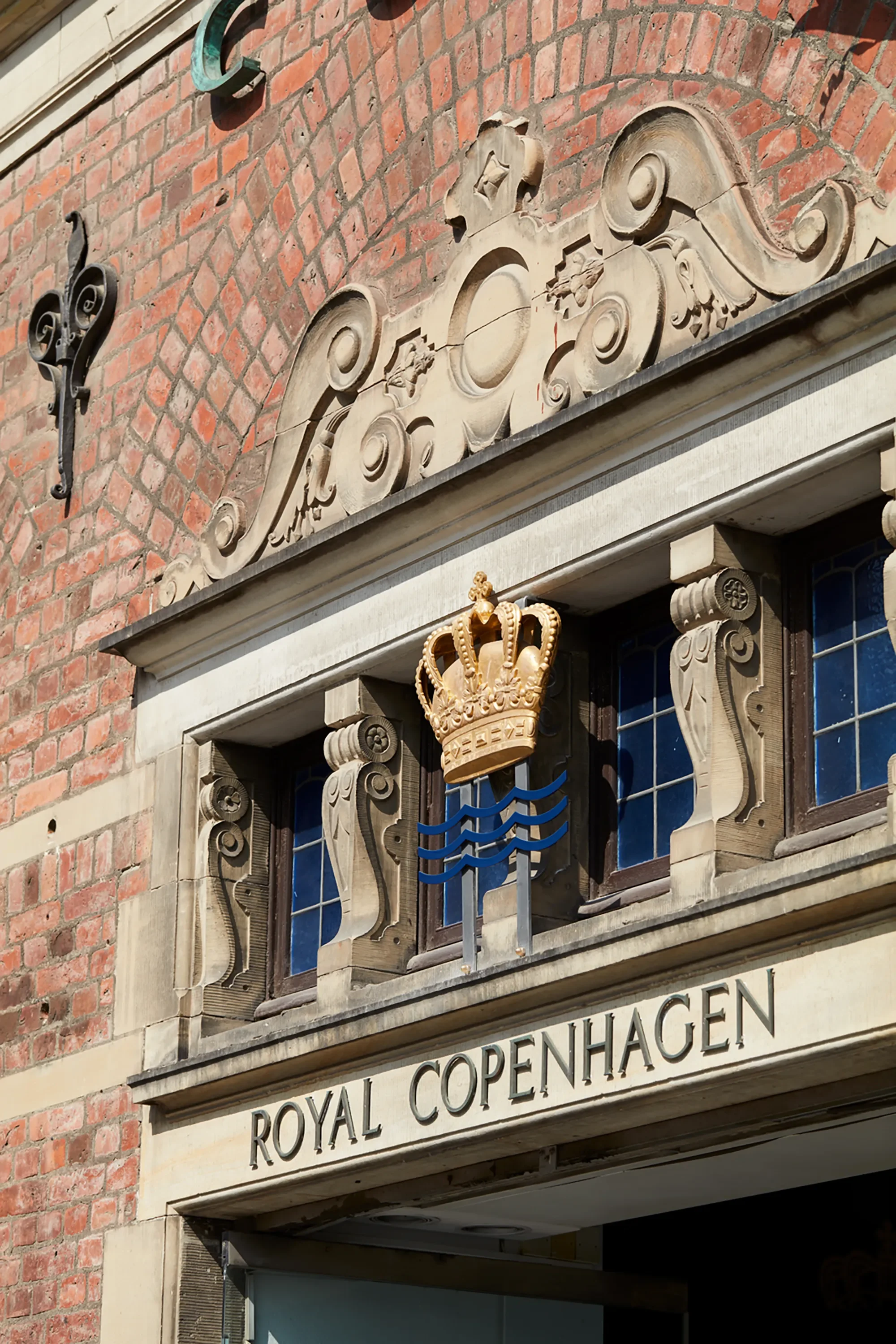 Close-up view of Royal Copenhagen's flagship store facade in Copenhagen, Denmark, featuring decorative stonework, a crown emblem, and part of the sign reading 'Royal Copenhagen' under a small part of an arched window with blue stained glass.
